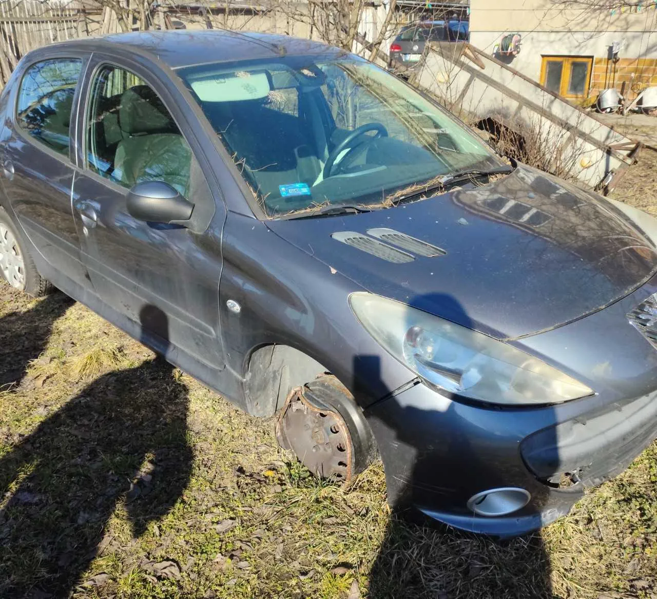 Ciemnoszary samochód osobowy z brakującym przednim lewym kołem, stojący na trawie z suchymi liśćmi. Na masce widoczne są gałązki i ślad po uszkodzeniu. W tle dom z żółtym oknem, suche krzewy i inny samochód. Na przedniej szybie naklejka z nieczytelnym tekstem. Cień osoby fotografującej widoczny na trawie i nadwoziu.