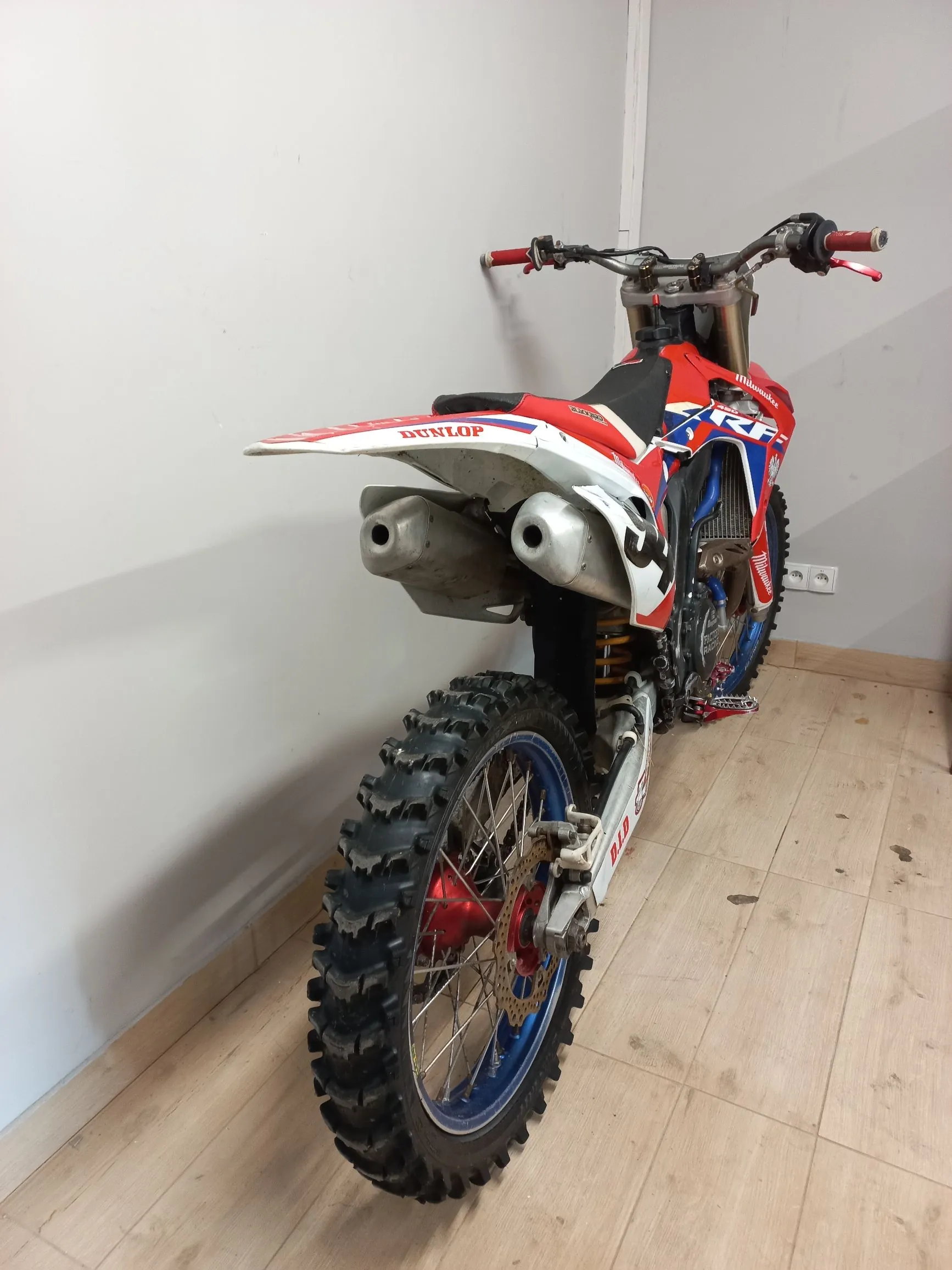 Motocykl crossowy w kolorach czerwonym, białym i niebieskim, ustawiony bokiem do białej ściany na jasnej drewnianej podłodze. Widoczny jest tylny terenowy opona z grubym bieżnikiem, niebieska obręcz koła, czerwona piasta oraz srebrny wydech. Na nadwoziu widnieją napisy 'DUNLOP' oraz 'RF'.