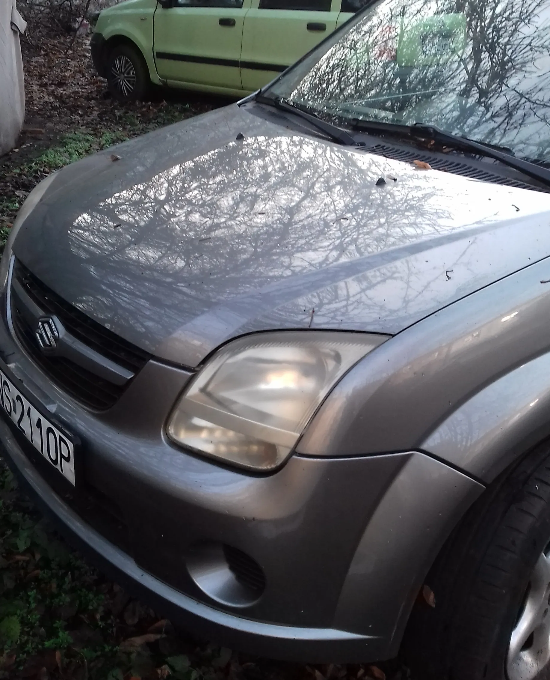 Zbliżenie na przód szarego samochodu marki Suzuki. Widoczna jest maska z odbiciem bezlistnych drzew, lewy reflektor oraz fragment przedniego zderzaka z częścią tablicy rejestracyjnej o treści 'S 2110P'. W tle widoczny jest fragment jasnozielonego samochodu zaparkowanego na trawiastym terenie.