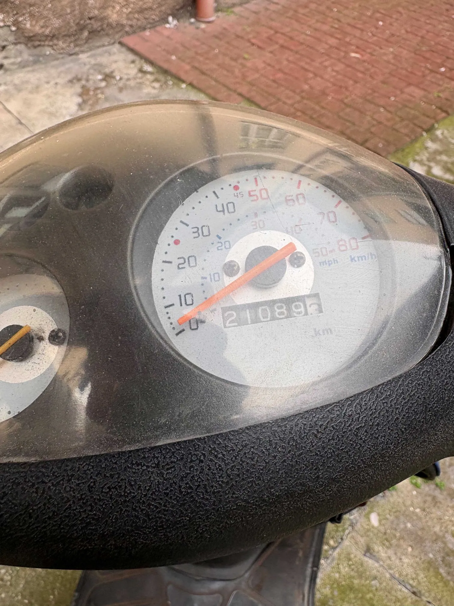 Zbliżenie na licznik prędkości skutera pod przezroczystą osłoną. Prędkościomierz wskazuje prędkość 0 km/h, a licznik kilometrów pokazuje wartość 21089 km. Skala prędkościomierza sięga 80 km/h. Wskazówka jest pomarańczowa.