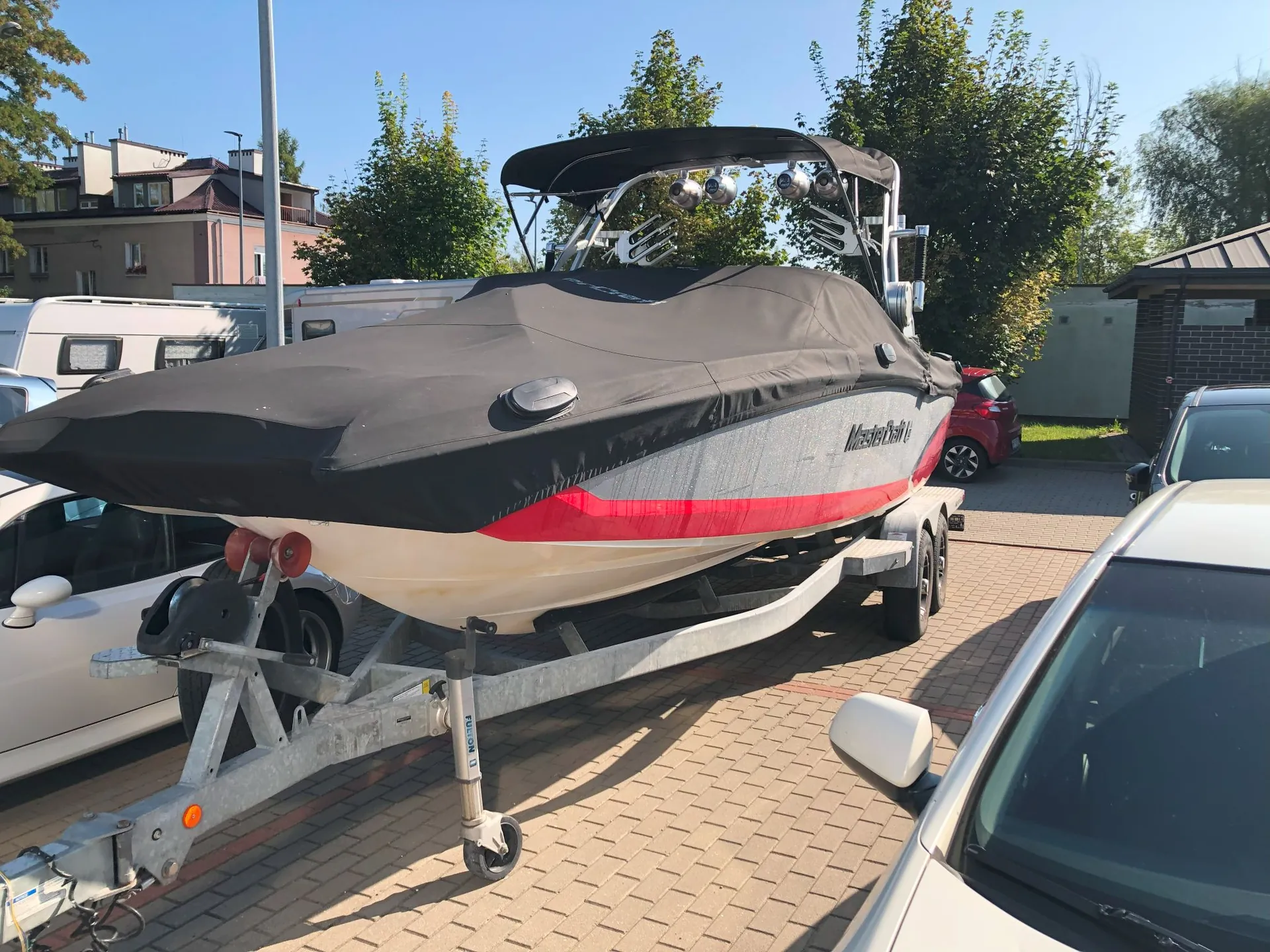Motorówka na przyczepie transportowej zaparkowana na kostce brukowej. Łódź jest przykryta czarną plandeką z czerwonym pasem u dołu i napisem 'MasterCraft'. W tle widoczne są drzewa, budynki mieszkalne oraz zaparkowane samochody, w tym biały samochód po lewej i prawej stronie oraz czerwony samochód w oddali.