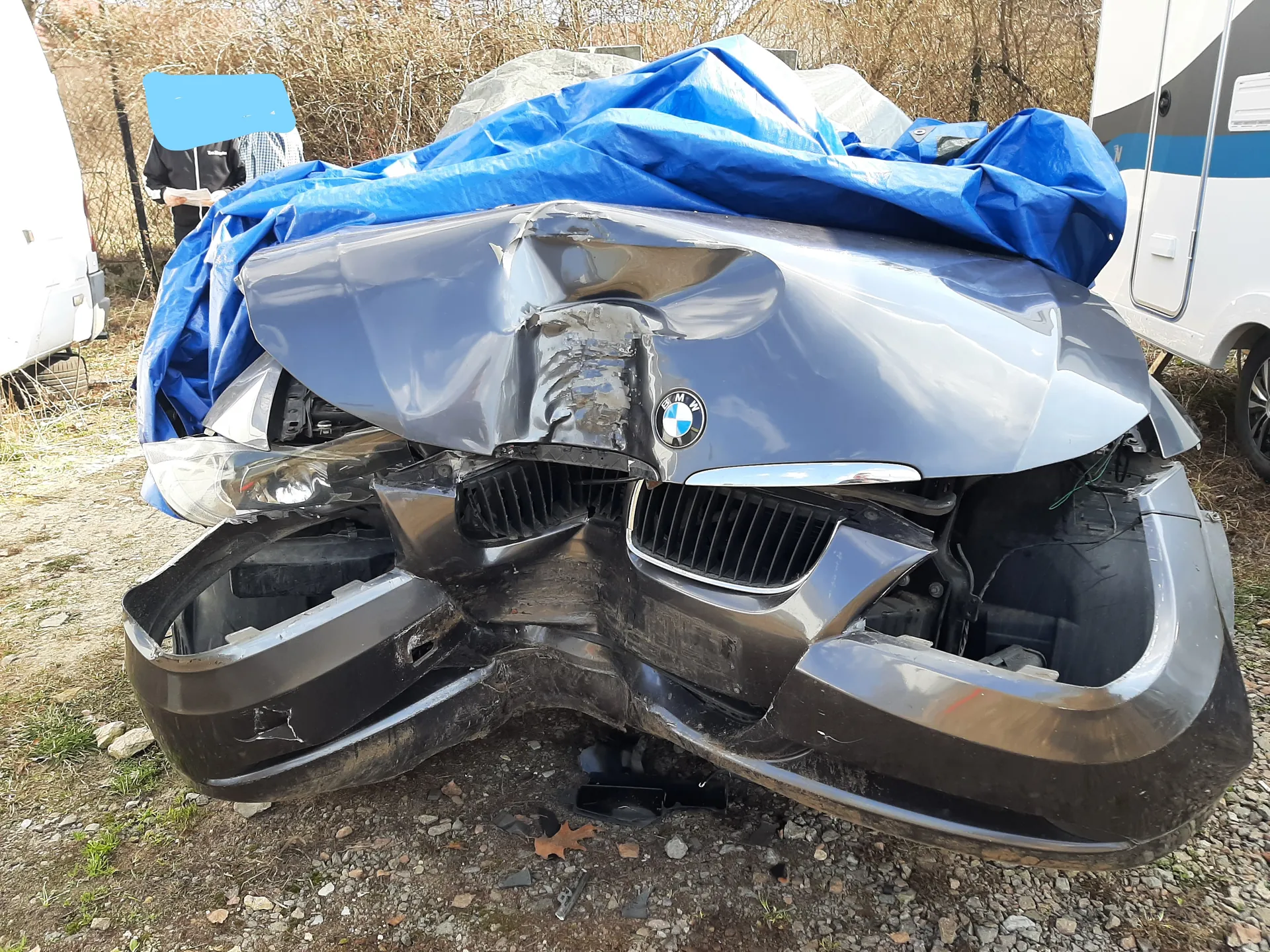 Zniszczony przód srebrnego samochodu marki BMW z widocznym logo na masce. Przednia część pojazdu jest silnie zdeformowana, z uszkodzonymi reflektorami i rozbitą atrapą chłodnicy. Na samochodzie leży niebieska folia. W tle widoczne są inne pojazdy: biały van po lewej stronie i biały przyczepa z niebieskimi pasami po prawej. Na tle suchych krzewów stoi osoba z zamazaną twarzą. Podłoże to żwirowa powierzchnia z fragmentami trawy i suchymi liśćmi.
