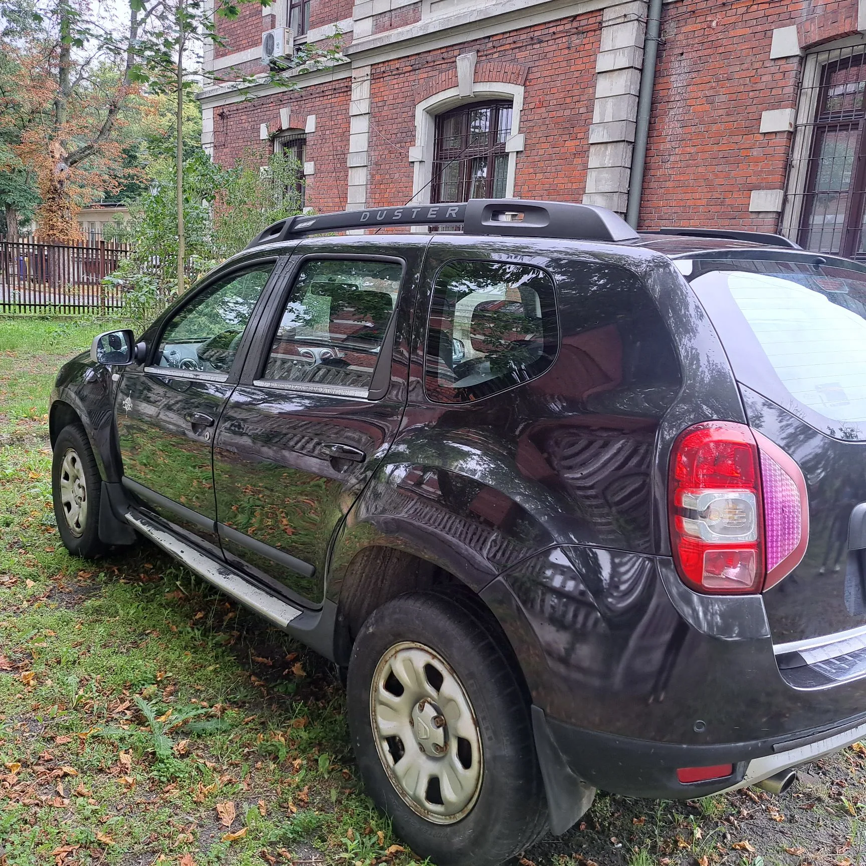 Czarny samochód marki Dacia Duster zaparkowany na trawie przed budynkiem z czerwonej cegły. Pojazd jest widoczny z profilu od strony pasażera, z widocznym napisem 'DUSTER' na relingu dachowym. W tle znajduje się ceglany budynek z oknami i fragment ogrodzenia.