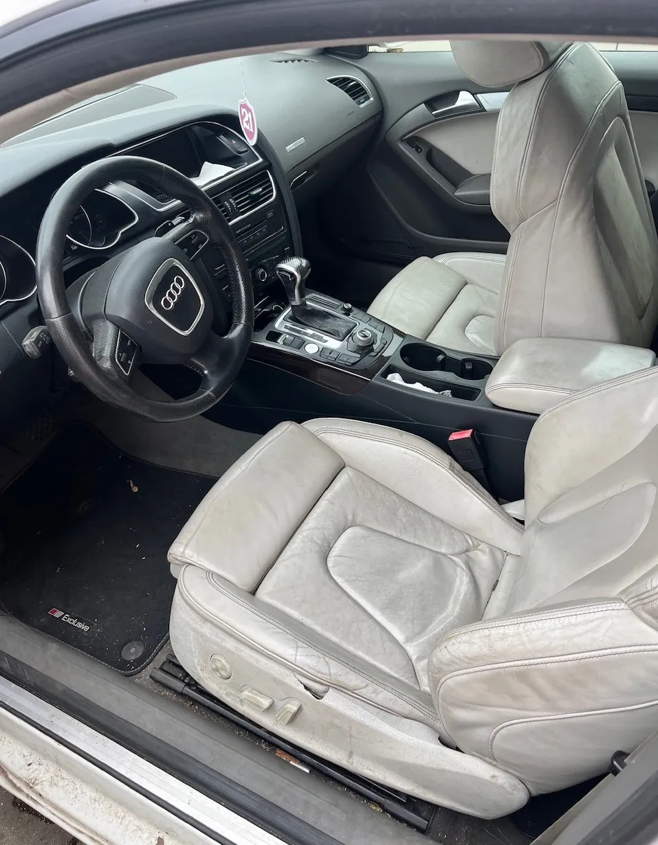 Wnętrze samochodu z widokiem na jasnoszare, skórzane fotele, deskę rozdzielczą oraz kierownicę z logo Audi. Widoczna jest automatyczna skrzynia biegów, konsola środkowa oraz dywanik z napisem 'Exclusive'.