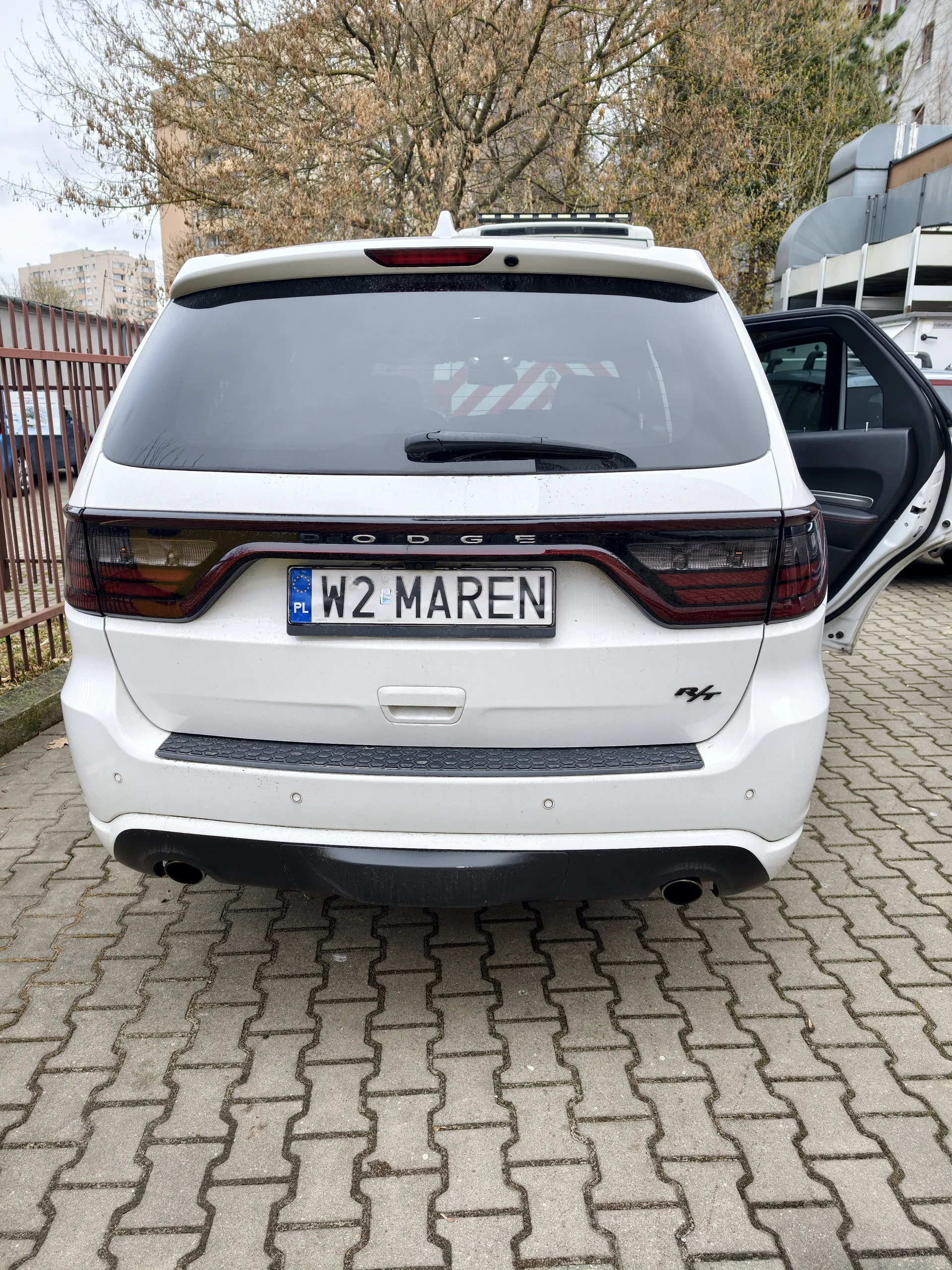 Tył białego samochodu marki Dodge z tablicą rejestracyjną W2 MAREN (PL). Drzwi po prawej stronie są otwarte. Tło: drzewa, budynki i ogrodzenie. Samochód stoi na brukowanej powierzchni.