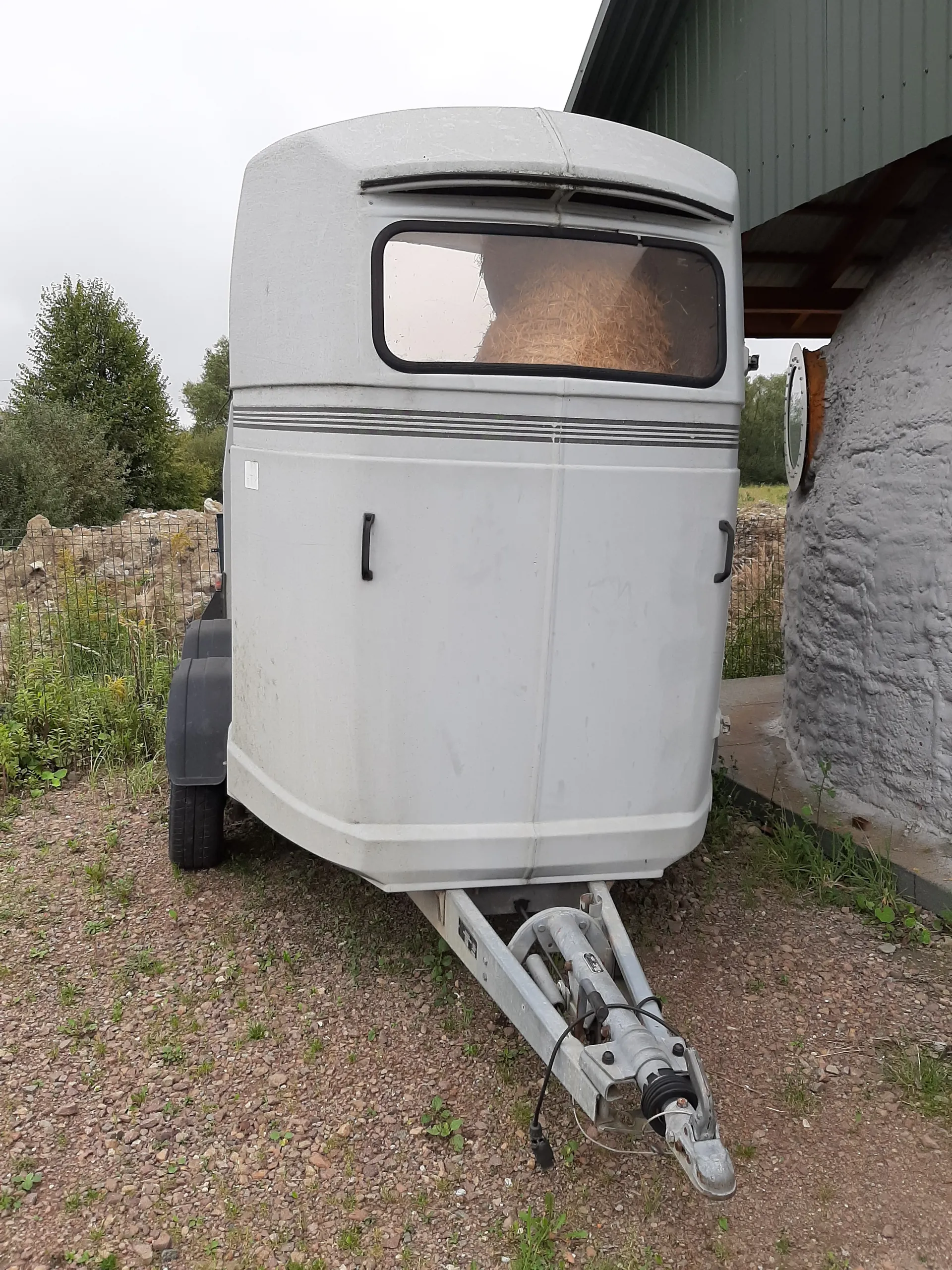 Szara przyczepa z oknem, przez które widać siano, stoi na żwirowym podwórku. Przyczepa posiada czarne uchwyty, elementy zaczepu i koła. W tle widoczny budynek z metalowym dachem, betonowa ściana, drzewa oraz siatka ogrodzeniowa.