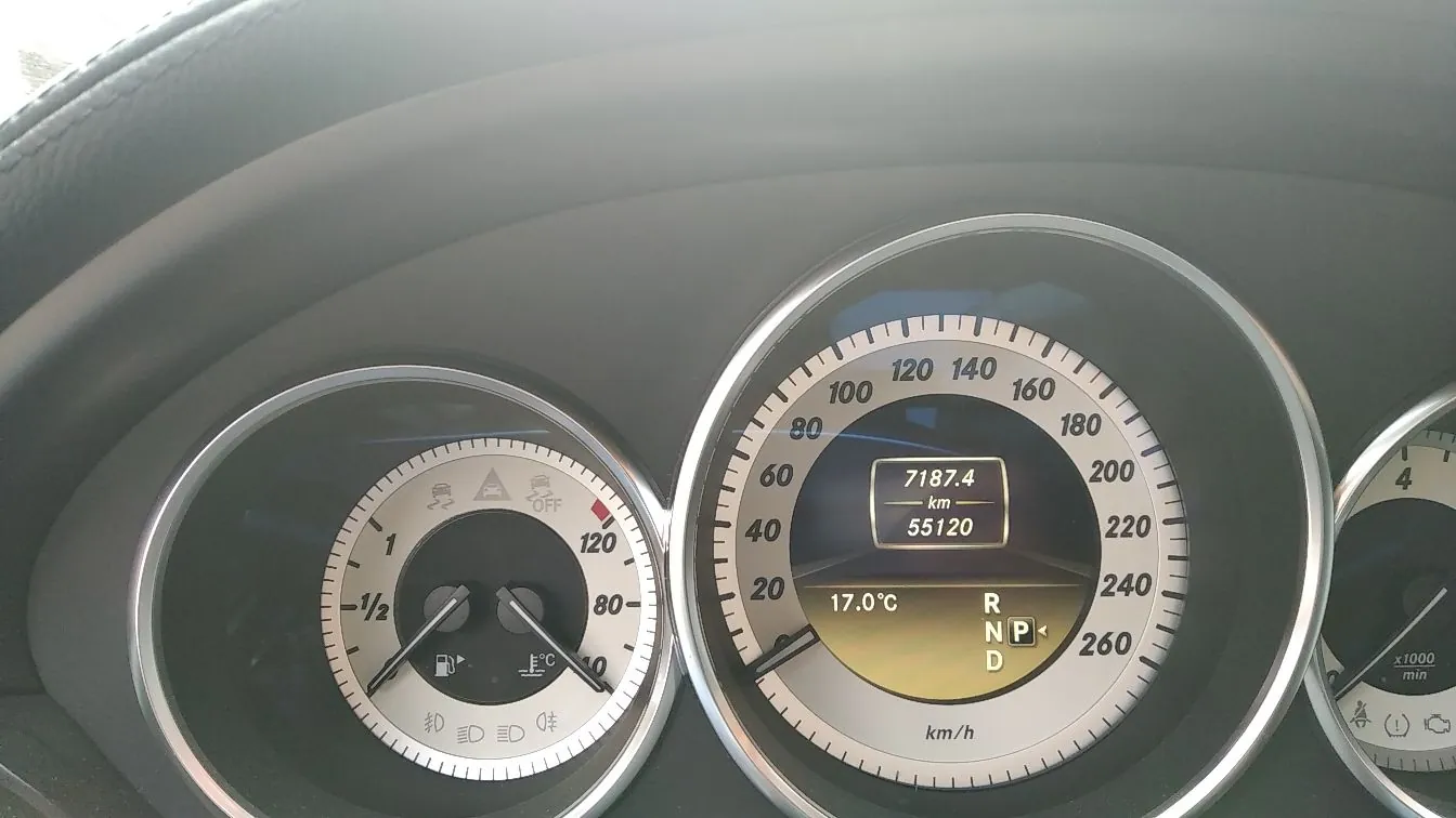 Zbliżenie na deskę rozdzielczą samochodu z trzema okrągłymi wskaźnikami. Środkowy wskaźnik to prędkościomierz z cyframi od 20 do 260 km/h, w którym znajduje się cyfrowy wyświetlacz pokazujący przebieg 55120 km, dystans 7187.4 km, temperaturę 17.0°C oraz aktualny bieg 'D'. Po lewej stronie znajduje się wskaźnik poziomu paliwa i temperatury silnika, a po prawej fragment obrotomierza.