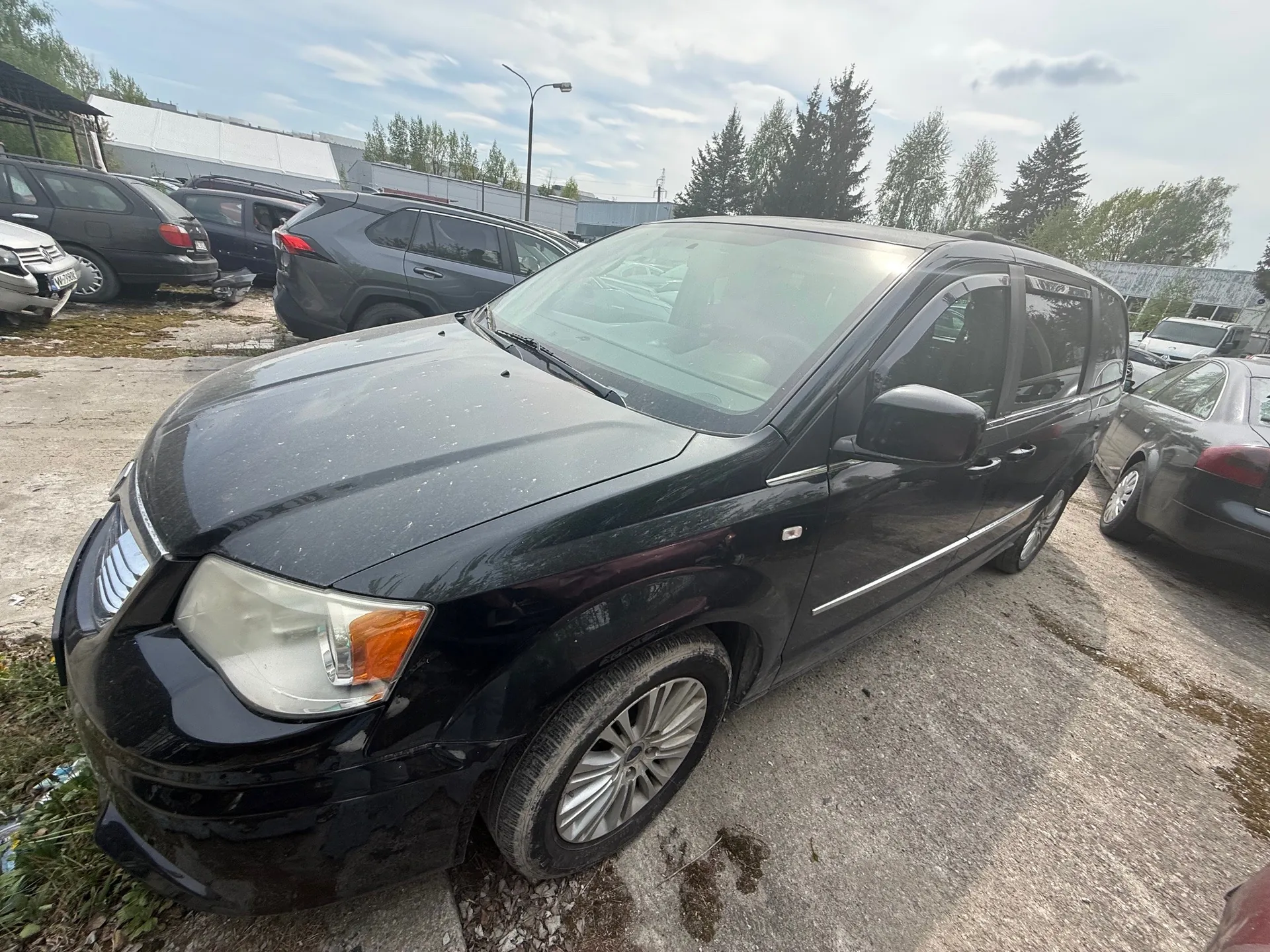 Czarny minivan zaparkowany na betonowym placu parkingowym. W tle inne samochody, w tym szary SUV i ciemne sedany, drzewa, przemysłowe budynki oraz słup latarni ulicznej. Niebo częściowo zachmurzone.