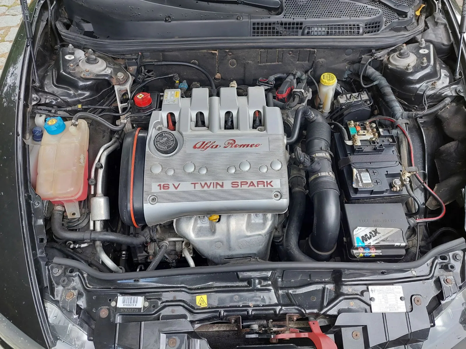 Widok z góry na komorę silnika samochodu marki Alfa Romeo. Centralnie znajduje się srebrna pokrywa silnika z czerwonymi napisami „Alfa Romeo” oraz „16 V TWIN SPARK”. Po lewej stronie widoczny jest przezroczysty zbiornik z płynem spryskiwaczami z niebieską zakrętką. Po prawej stronie znajduje się akumulator z widocznymi zaciskami oraz czarne rury dolotowe powietrza. Całość jest osadzona w czarnej ramie nadwozia pojazdu.