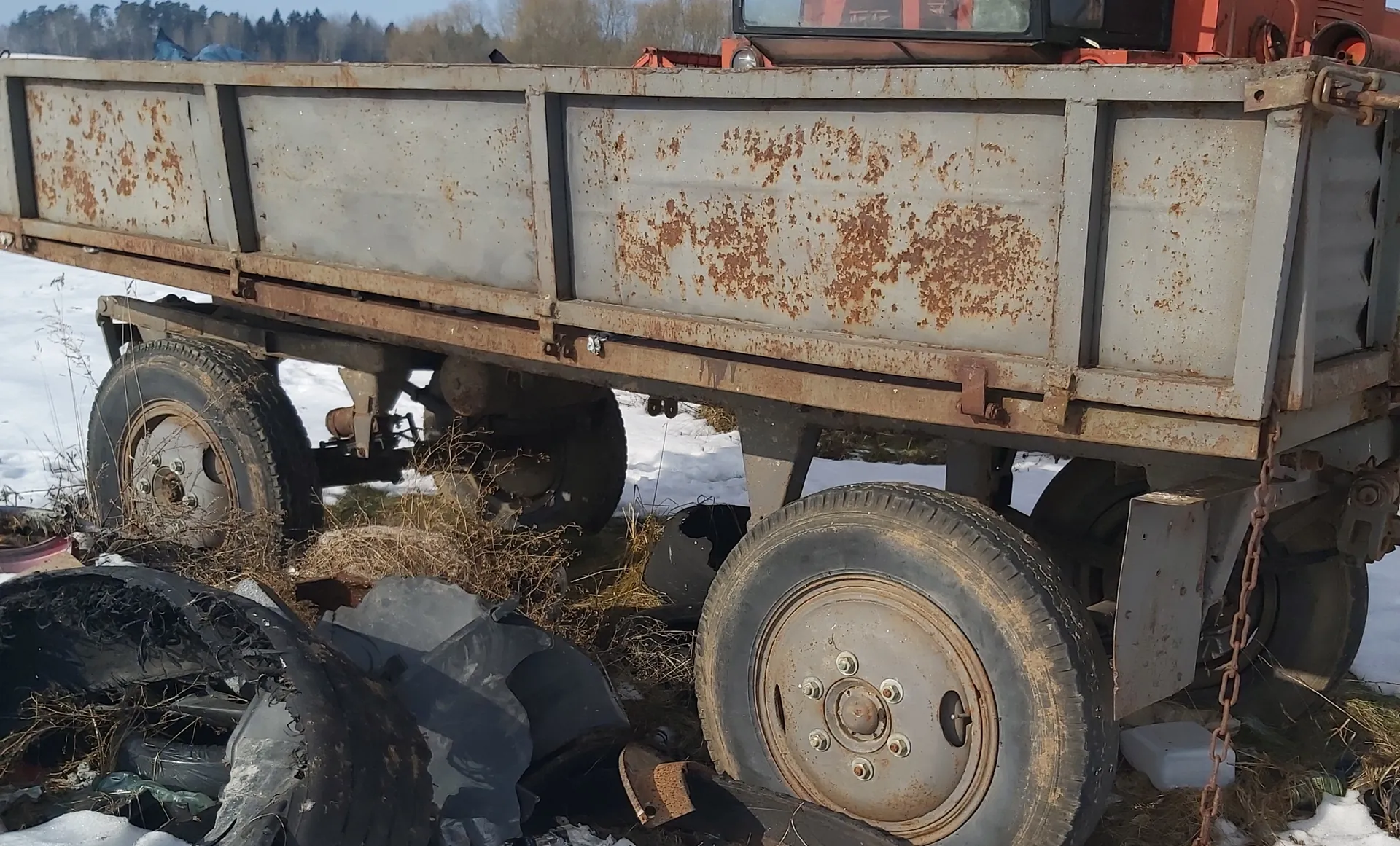 Zardzewiały metalowy zbiornik przyczepy ciężarowej z widocznymi plamami rdzy, stojący na śniegu i suchych trawach. Wokół rozrzucone są zużyte opony, niektóre uszkodzone. W tle drzewa i jasne niebo. Część pomarańczowego pojazdu widoczna w górnej części obrazu.