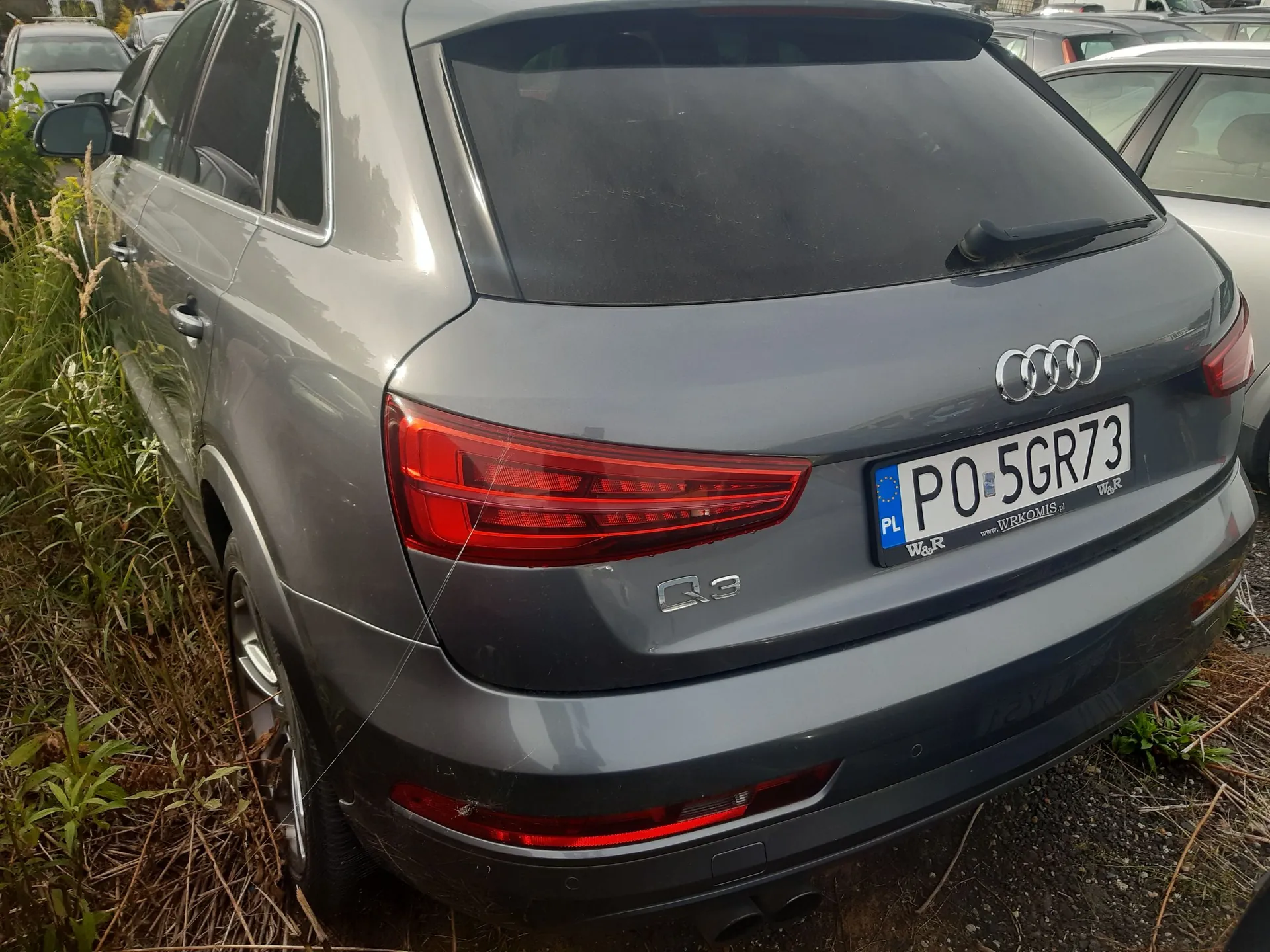 Tylna część szarego samochodu marki Audi model Q3. Widoczny jest znak rejestracyjny PO 5GR73 oraz logo marki Audi. Samochód zaparkowany jest na poboczu obok zielonej roślinności.