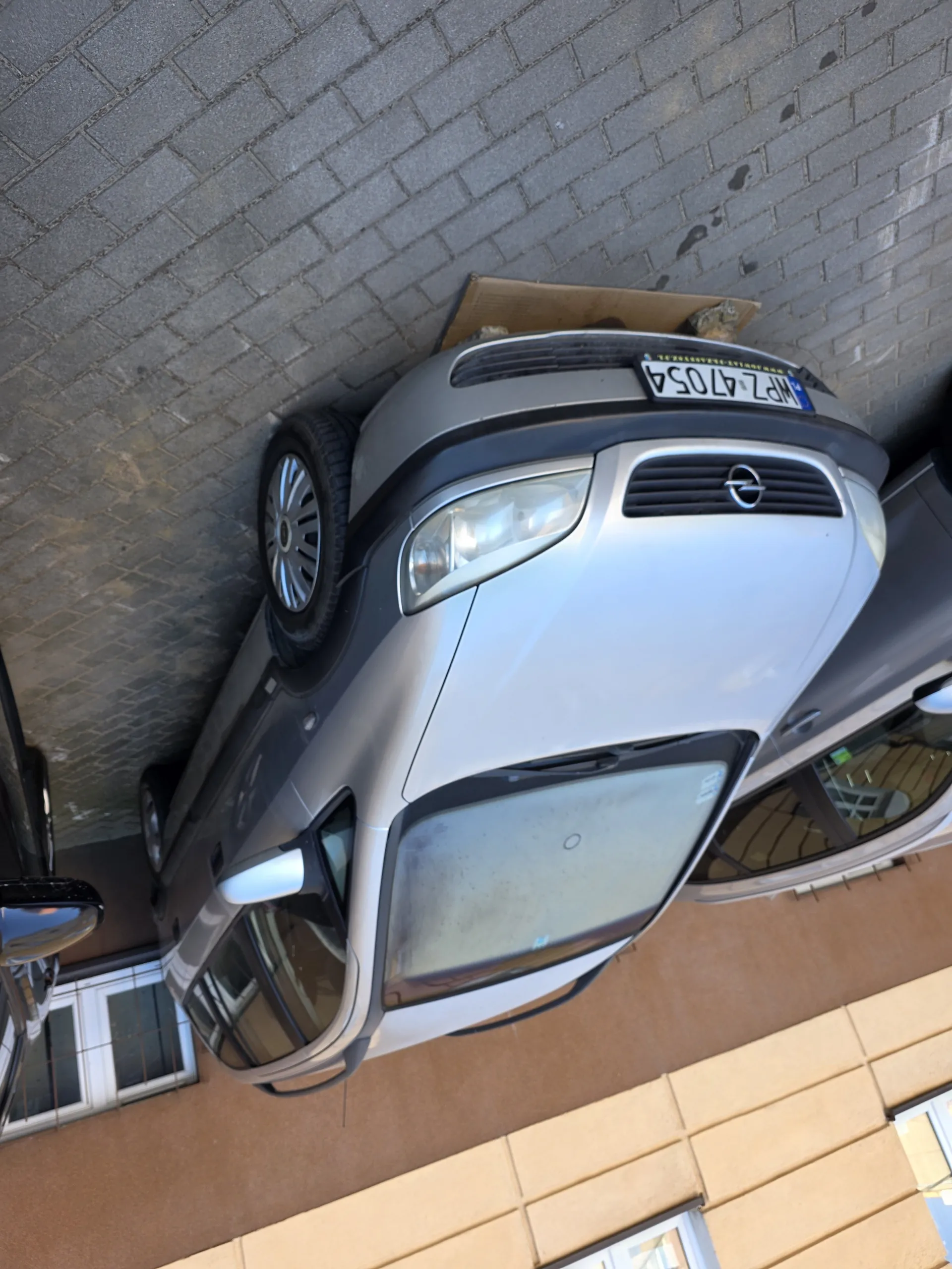 Srebrny samochód marki Opel zaparkowany na brukowanej powierzchni. Widoczna tablica rejestracyjna WPZ 47054. Za nim częściowo widoczny inny samochód. W tle budynek z brązową ścianą i oknami.