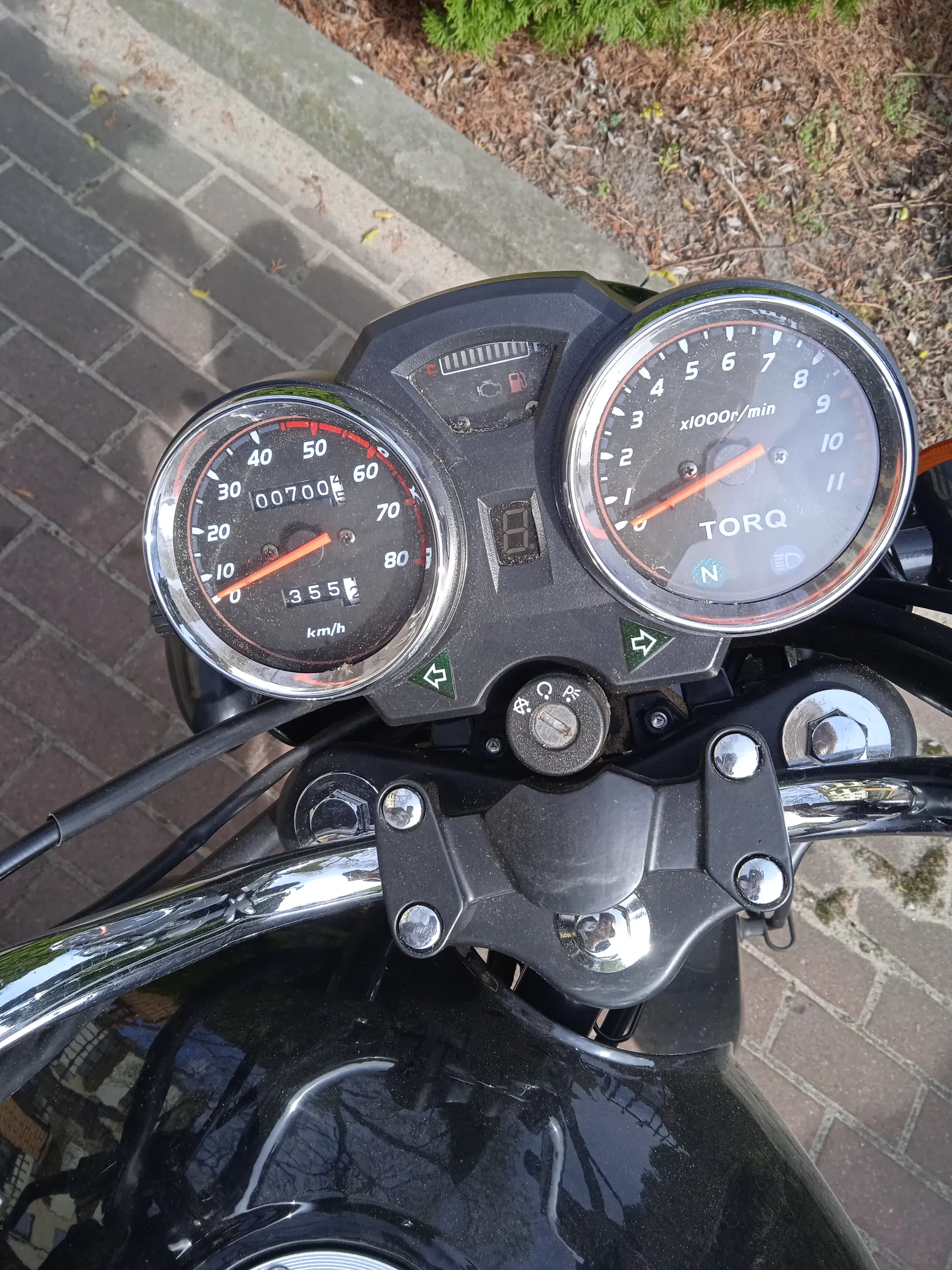 Zbliżenie na deski rozdzielcze motocykla marki TORQ. Po lewej stronie znajduje się prędkościomierz z zakresem 0-80 km/h, pokazujący 0 km/h, z licznikiem przebiegu 00700 i licznikiem dziennym 355. Po prawej stronie tachometr z zakresem 0-11 (x1000r/min), z czerwoną igłą wskazującą na 0. W centralnej części cyfrowy wyświetlacz z cyfrą 8 oraz wskaźniki paliwa i neutrala. Widoczne są również kierownica z metalowymi elementami i tłem z brukowanego chodnika oraz zieleni.