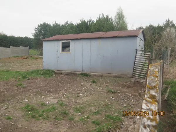 Zdjęcie przedstawia prosty, prostokątny budynek gospodarczy o jasnoniebieskich ścianach i brązowym, lekko spadzistym dachu. W ścianie znajduje się jedno małe, kwadratowe okno. Budynek stoi na nieutwardzonym, piaszczystym terenie z kępami trawy. Po prawej stronie widoczna jest metalowa brama oraz fragment niskiego, kamiennego murka. W tle znajduje się gęsty las iglasty pod zachmurzonym niebem. W prawym dolnym rogu widnieje data i godzina: 15/04/2026 10:00.