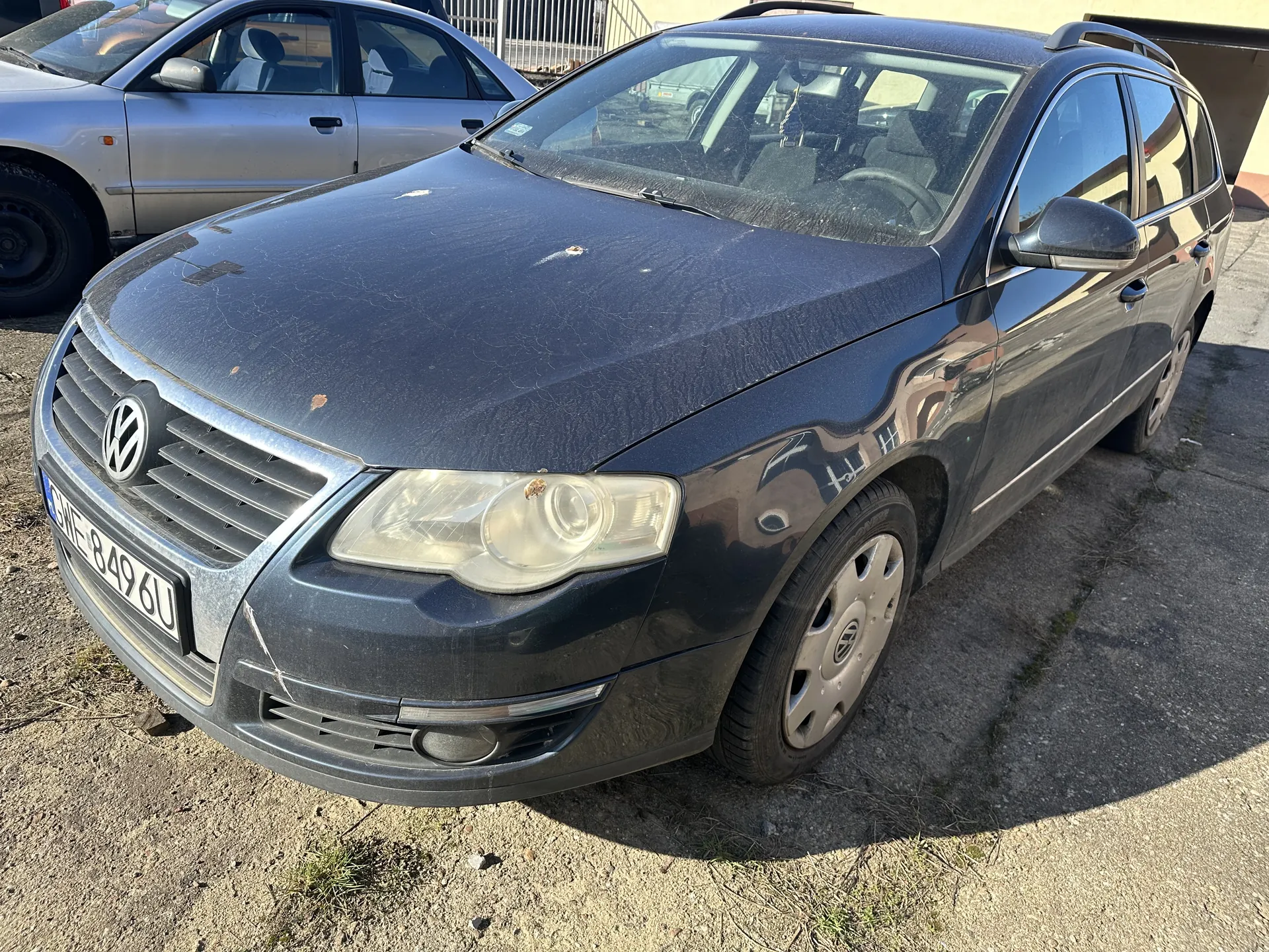 Ciemnoszary samochód osobowy marki Volkswagen, typu kombi, zaparkowany na nierównym podłożu z drobnymi kamieniami i trawą. Na przodzie widoczna tablica rejestracyjna GWE 849 6U, logo VW na atrapie chłodnicy oraz reflektory. Samochód posiada dachowy bagażnik i lusterka boczne. W tle drugi srebrny samochód osobowy oraz fragment budynku z metalowym ogrodzeniem. Na karoserii widoczne są drobne uszkodzenia i zabrudzenia.