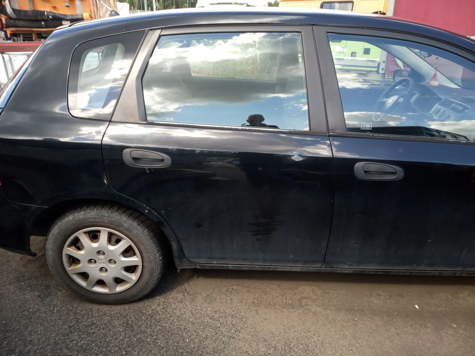 Boczny widok czarnego samochodu osobowego typu kombi. Na tylnych drzwiach pasażera widoczne jest wgniecenie oraz zarysowania lakieru. Samochód stoi na szutrowym podłożu.