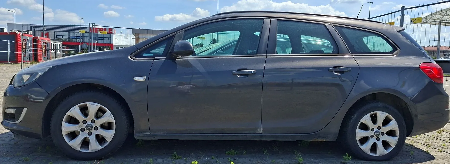 Profil ciemnoszarego samochodu typu kombi zaparkowanego na kostce brukowej. W tle widoczne są budynki przemysłowe, w tym obiekt z czerwono-białym logo 'MERLE', oraz ogrodzenie z siatki pod jasnym, lekko zachmurzonym niebem.