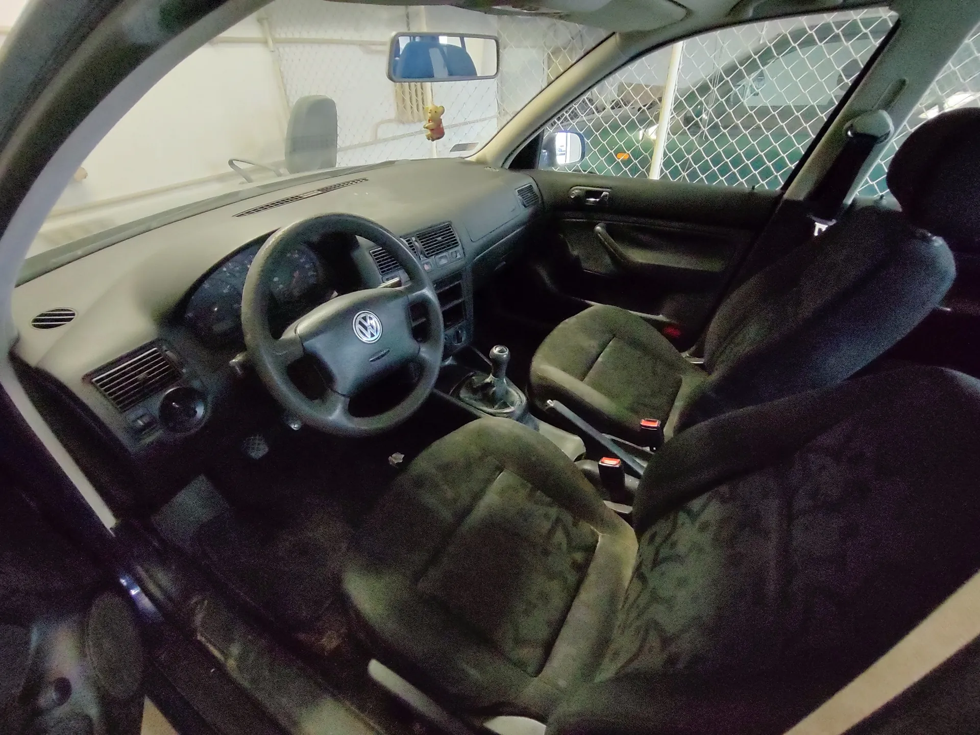 Wnętrze samochodu marki Volkswagen z widocznym kierownicą z logo VW, deska rozdzielcza w kolorze beżowym, czarne siedzenia, dźwignia zmiany biegów oraz lusterko wsteczne. Przez przednią szybę widoczny jest ogrodzenie siatkowe i inny pojazd. W tle na szybie wisząca zabawka w kształcie misia.