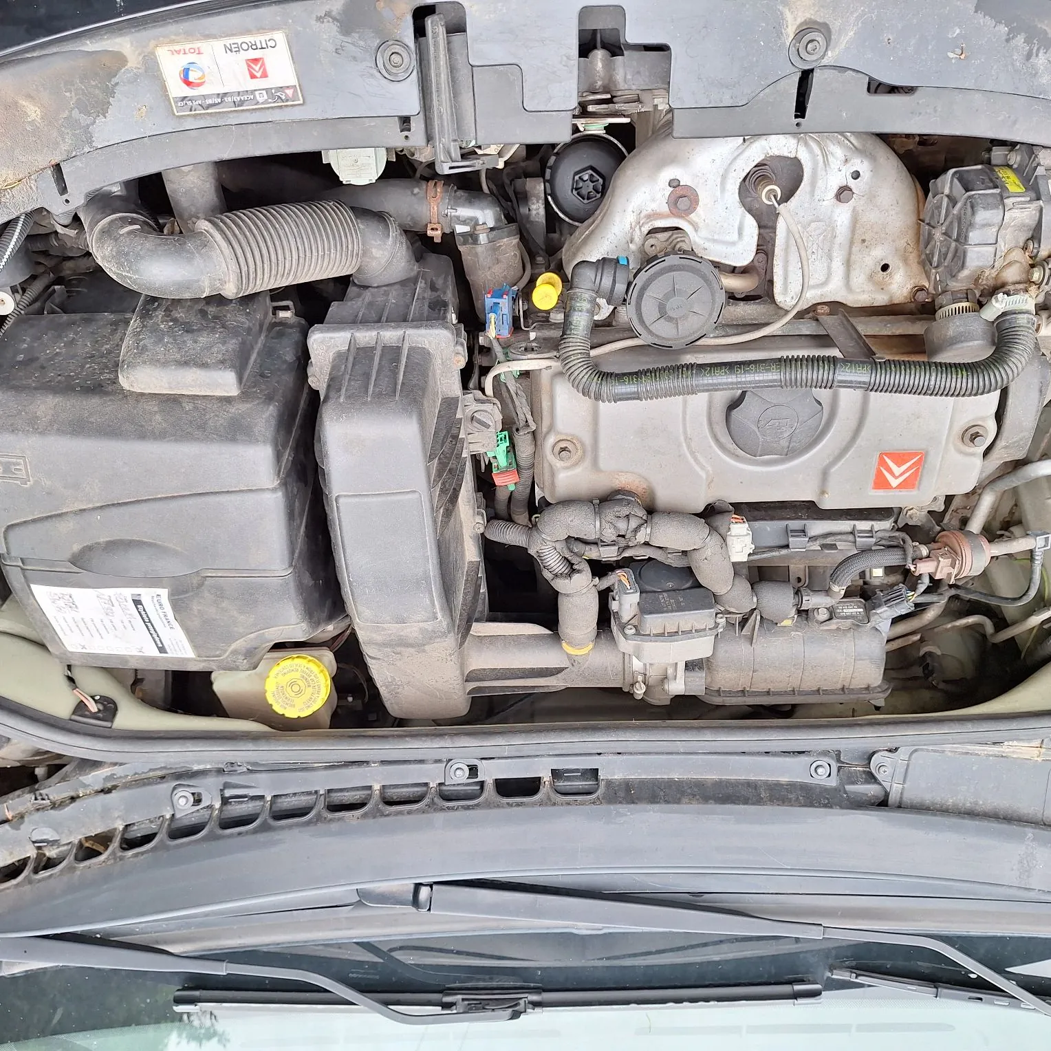 Zdjęcie przedstawiające komorę silnika samochodu marki Citroën. Widoczne są elementy mechaniczne, w tym obudowa filtra powietrza, przewody, akumulator oraz metalowe części silnika. W górnej części znajduje się naklejka z napisem 'CITROEN TOTAL'. Obraz jest zorientowany tak, że przód samochodu znajduje się po lewej stronie, a podszybie na dole.