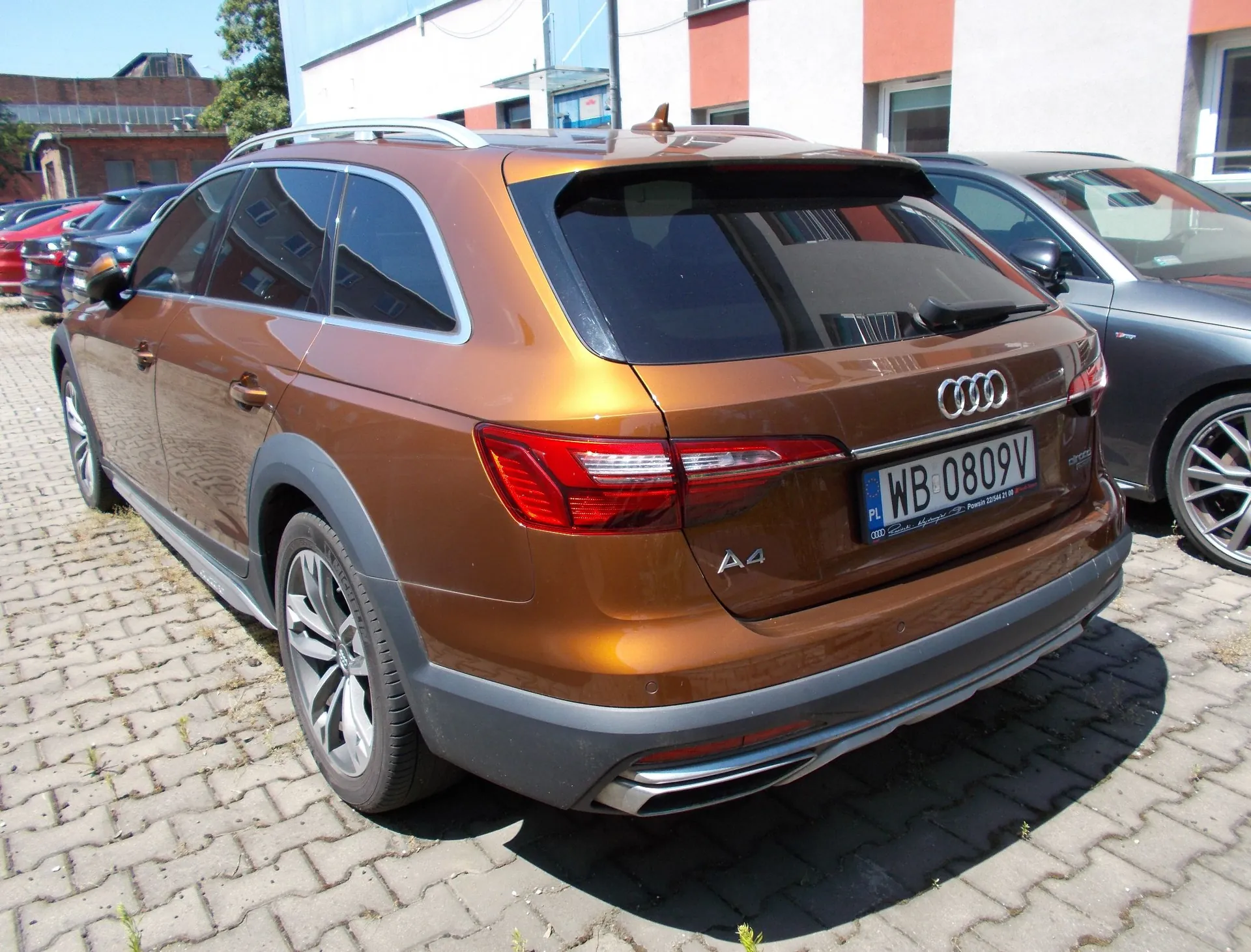 Brązowy samochód marki Audi A4 Allroad zaparkowany na kostce brukowej. Widoczny jest tył i lewy bok pojazdu. Samochód posiada tablicę rejestracyjną o numerze WB 0809V. W tle widoczne są inne zaparkowane samochody oraz fragmenty budynków.