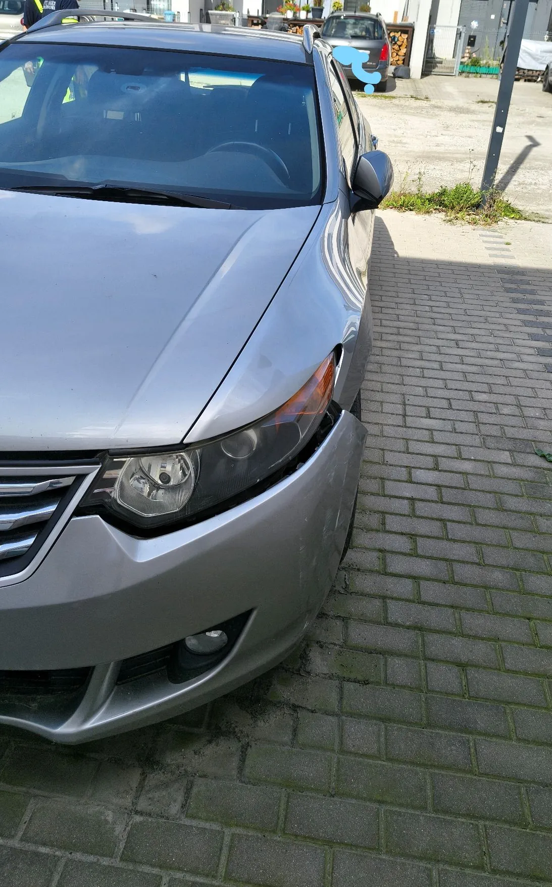 Srebrny samochód osobowy zaparkowany na kostce brukowej. Widoczny jest przód pojazdu z reflektorem, maską i fragmentem zderzaka. W tle widać inny samochód oraz fragmenty budynków.