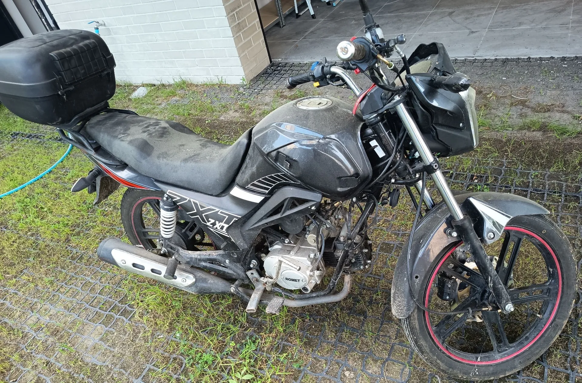 Czarny motocykl marki Romet z zamontowanym czarnym kufrem z tyłu, zaparkowany na trawiastym podłożu wzmocnionym plastikową kratką. Motocykl jest zakurzony, posiada czerwone detale na obręczach kół oraz napis 'ZX1' na boku. W tle widoczna jest biała ściana z cegieł i fragment betonowego podjazdu.