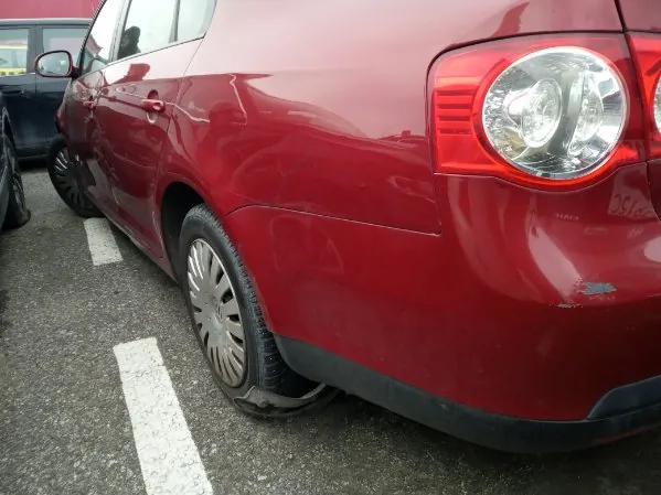 Czerwony samochód zaparkowany na asfaltowym parkingu z białymi liniami. Widoczna tylna część pojazdu, w tym tylne światło, koło z płaską oponą oraz fragment sąsiedniego czarnego samochodu po lewej stronie. Na tle asfaltu i białych linii parkingu.