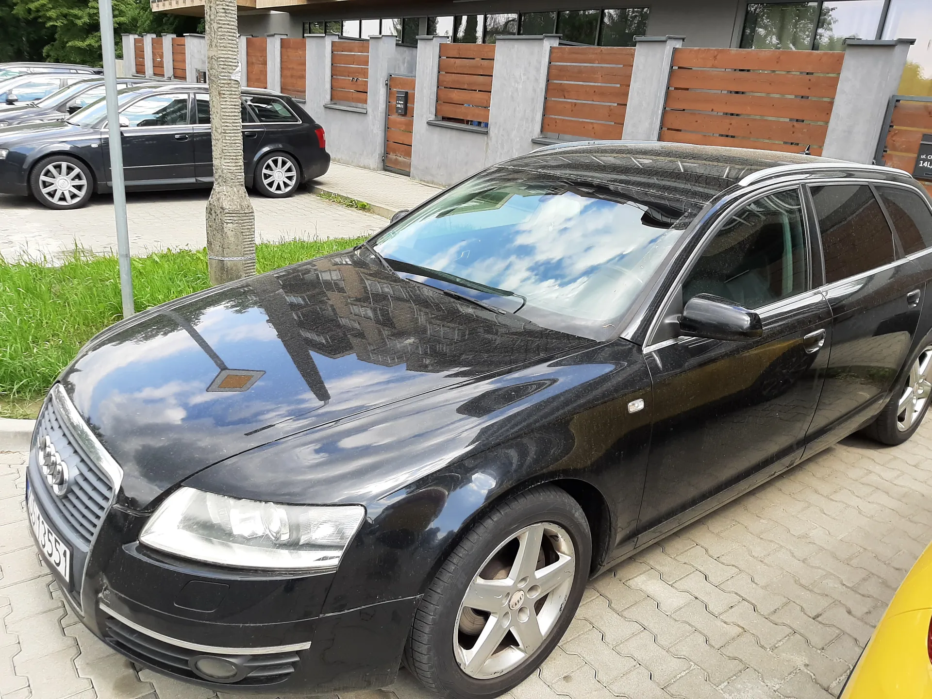 Czarny samochód Audi typu kombi zaparkowany na brukowanej powierzchni. Widoczna tablica rejestracyjna WU13551. W tle drugi czarny samochód, budynek z betonowymi filarami i drewnianym ogrodzeniem, drzewo oraz zielona trawa. Na masce odbicie nieba z chmurami i budynku.