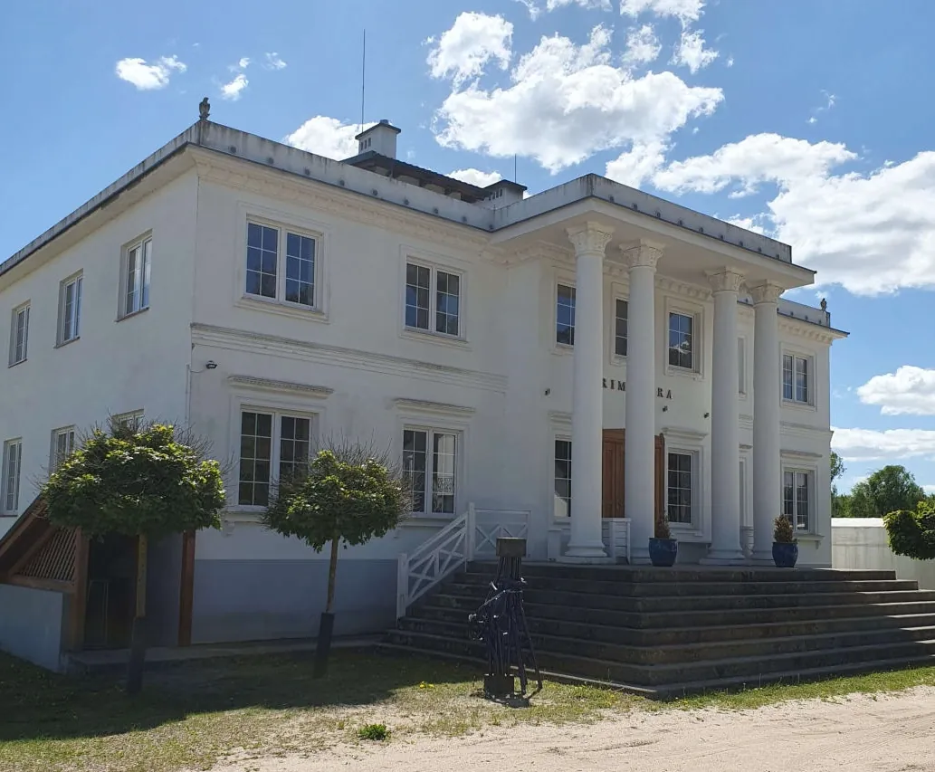 Biały dwupiętrowy budynek z kolumnami na fasadzie, schodami prowadzącymi do wejścia, oknami i drzewami na trawniku. Na budynku widoczny fragment tekstu 'RIM RA'. Tło: błękitne niebo z chmurami.
