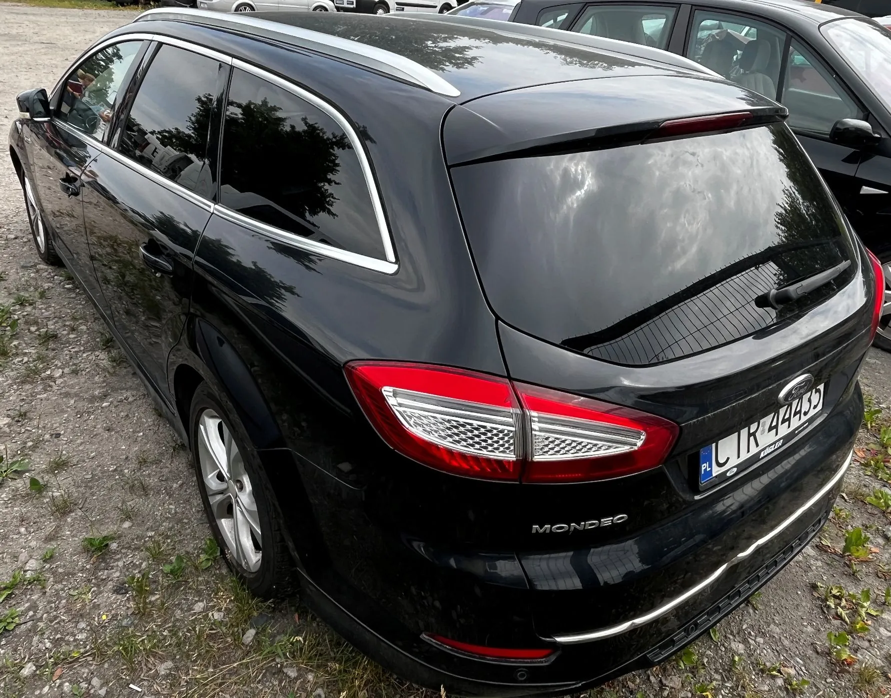 Czarny samochód marki Ford Mondeo w wersji kombi, zaparkowany na żwirowym podłożu. Widok z tyłu i z boku. Pojazd posiada srebrne relingi dachowe, chromowane obramowania szyb oraz polską tablicę rejestracyjną o numerze CTR 44433. Na klapie bagażnika widnieje napis MONDEO oraz logo Forda.