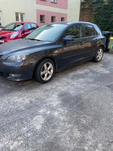 Ciemnoszary samochód osobowy typu hatchback zaparkowany na betonowej powierzchni. W tle czerwony samochód, budynek z różowymi i beżowymi ścianami oraz oknami, a także zielone drzewa. Na pierwszym planie widoczna nierówna, szara nawierzchnia.