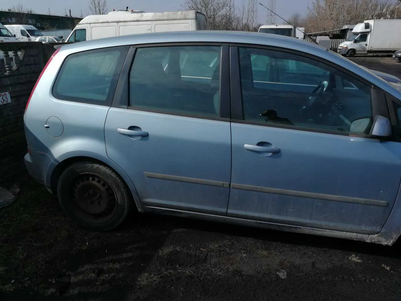 Niebieski samochód typu kombi zaparkowany na brudnej powierzchni. Widoczne są cztery drzwi, przyciemniane szyby, brak felgi na przednim lewym kole oraz zabrudzenia na dolnej części nadwozia. W tle: białe furgony, ciężarówka, ceglany mur z tabliczką z numerem 33 oraz drzewa bez liści. Niebo jest jasne.