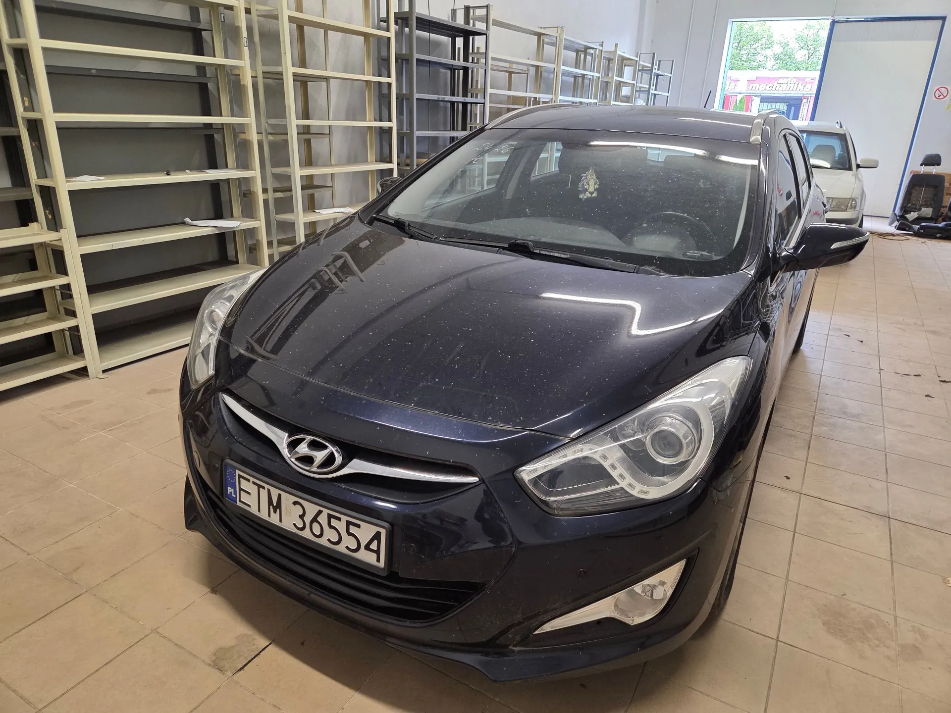 Granatowy samochód marki Hyundai zaparkowany wewnątrz pomieszczenia przypominającego magazyn lub warsztat. Auto jest zakurzone, widoczna jest tablica rejestracyjna o numerze ETM 36554. W tle znajdują się metalowe regały magazynowe oraz fragment innego, jasnego samochodu. Po prawej stronie widoczne jest wejście do innego pomieszczenia z napisem 'mechanika'.