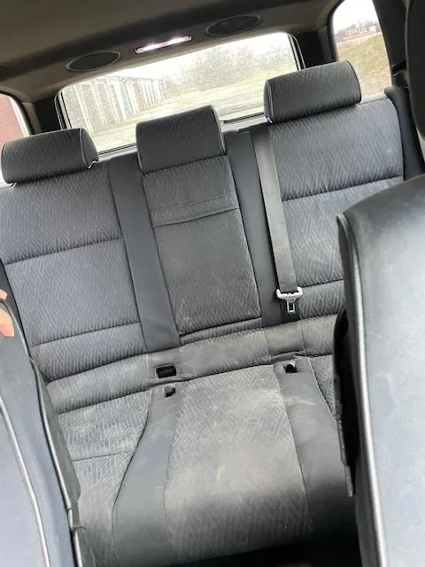 Wnętrze samochodu z widokiem na tylne siedzenia. Siedzenia są szare, tapicerowane tkaniną z geometrycznym wzorem, z trzema zagłówkami i pasami bezpieczeństwa. Widoczne są ślady zużycia i zabrudzenia na siedzeniach. Przez tylne okno widać zewnętrzny krajobraz z budynkami i drzewami. Wewnątrz samochodu widoczne są również elementy sufitu, w tym lampki oświetleniowe.