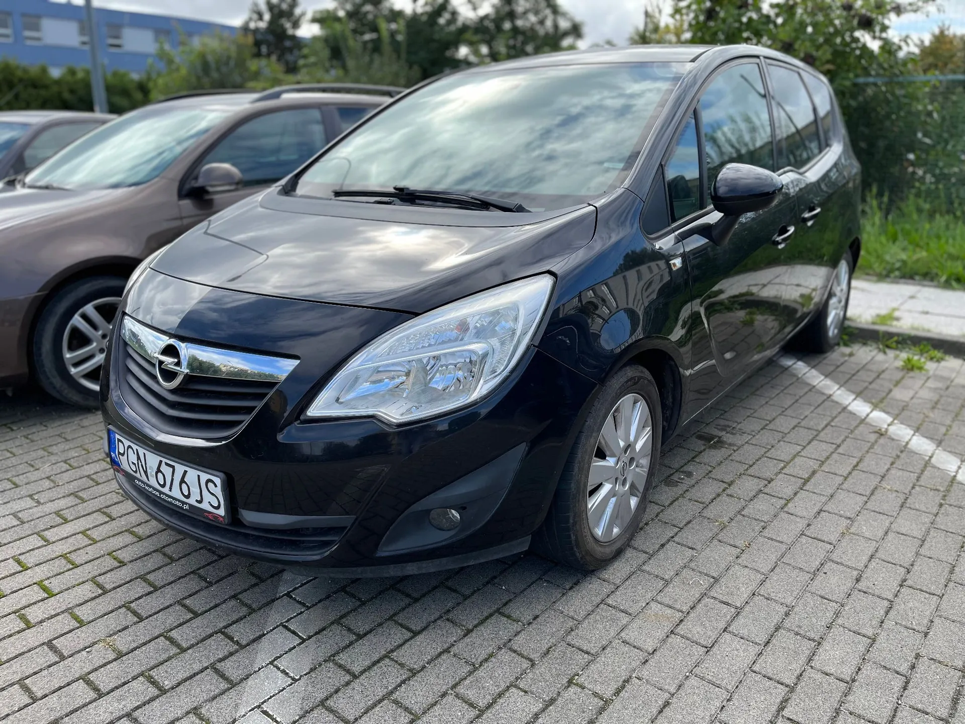 Czarny samochód marki Opel zaparkowany na kostce brukowej. Pojazd jest ustawiony pod kątem, widoczny z przodu i z boku. Na przednim zderzaku znajduje się polska tablica rejestracyjna o numerze PGN 676JS. W tle widoczne są inne zaparkowane samochody oraz zielone krzewy i fragment budynku.