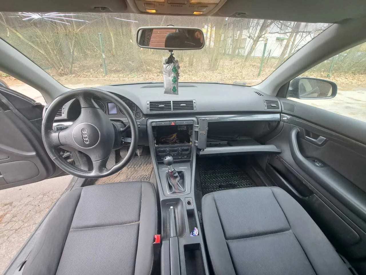 Wnętrze samochodu marki Audi z widocznym kierownicą z logo Audi, szarym deski rozdzielczej i siedzeniami. W centralnej konsoli brakuje urządzenia audio, widoczne są przewody. Ręczny hamulec i dźwignia zmiany biegów znajdują się na środku. Drzwi po stronie kierowcy są otwarte. Przez przednią szybę widać drzewa i budynek na zewnątrz. W lusterku wstecznym widoczny jest fragment wnętrza oraz wiszący na nim przedmiot z zielonymi symbolami.