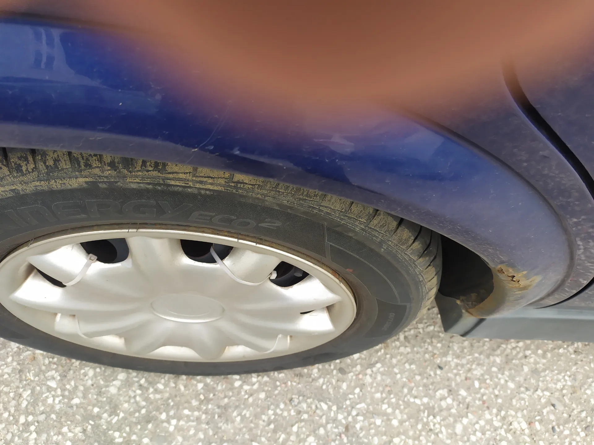 Zbliżenie na przednie koło niebieskiego samochodu z srebrnym felgiem. Opona ma widoczny napis "ENERGY ECO2" oraz zabrudzenia. Nadwozie samochodu w okolicy koła posiada ślady rdzy. Podłoga to asfalt z drobnymi kamieniami. Palcem częściowo zasłonięta górna część obrazu.