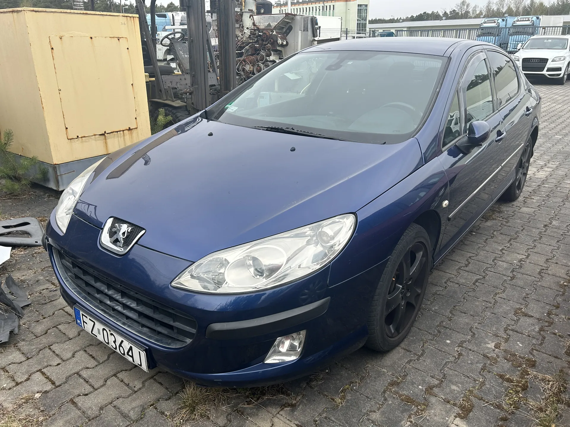 Niebieski samochód osobowy marki Peugeot z tablicą rejestracyjną PL FZ-0364J zaparkowany na brukowanej powierzchni z trawą między kamieniami. W tle widoczny żółty kontener, maszyny przemysłowe oraz biały samochód marki Audi. Niebo jest pochmurne.