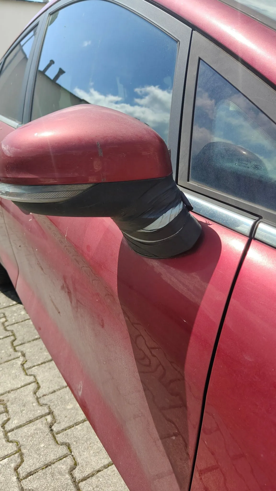Zbliżenie na czerwone lusterko boczne samochodu. Lusterko jest uszkodzone – dolna, czarna plastikowa obudowa jest pęknięta i częściowo oddzielona od górnej, czerwonej części. Karoseria samochodu wokół lusterka jest zabrudzona. W tle widoczne są szyby auta oraz fragment chodnika z kostki brukowej.
