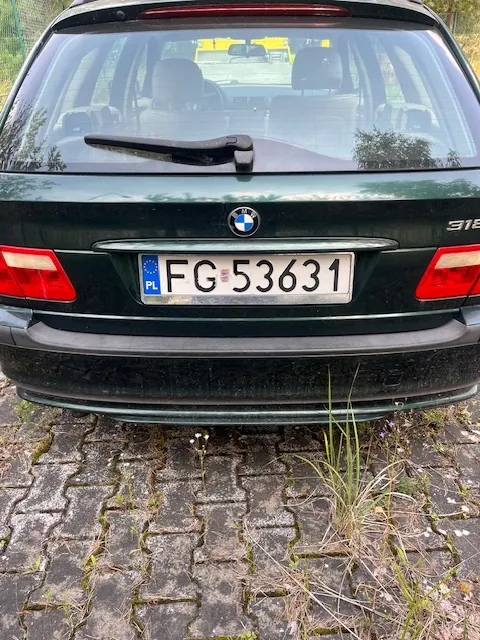 Tylny widok ciemnozielonego samochodu marki BMW. Na klapie bagażnika znajduje się logo BMW oraz oznaczenie modelu 318. Samochód posiada polską tablicę rejestracyjną o numerze FG 53631. Pojazd stoi na kostce brukowej z przerastającą trawą.
