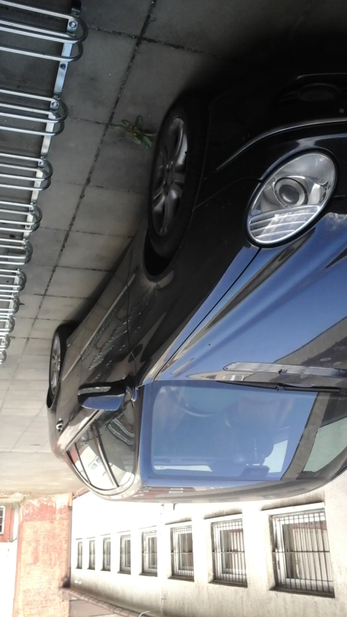 Czarny samochód osobowy zaparkowany przy betonowej ścianie z metalowymi uchwytami na rowery. Po lewej stronie widoczny budynek z białą elewacją i oknami z metalowymi kratami. Na betonowej powierzchni pod samochodem rośnie niewielka roślina. Samochód ma przyciemniane szyby i srebrne felgi.