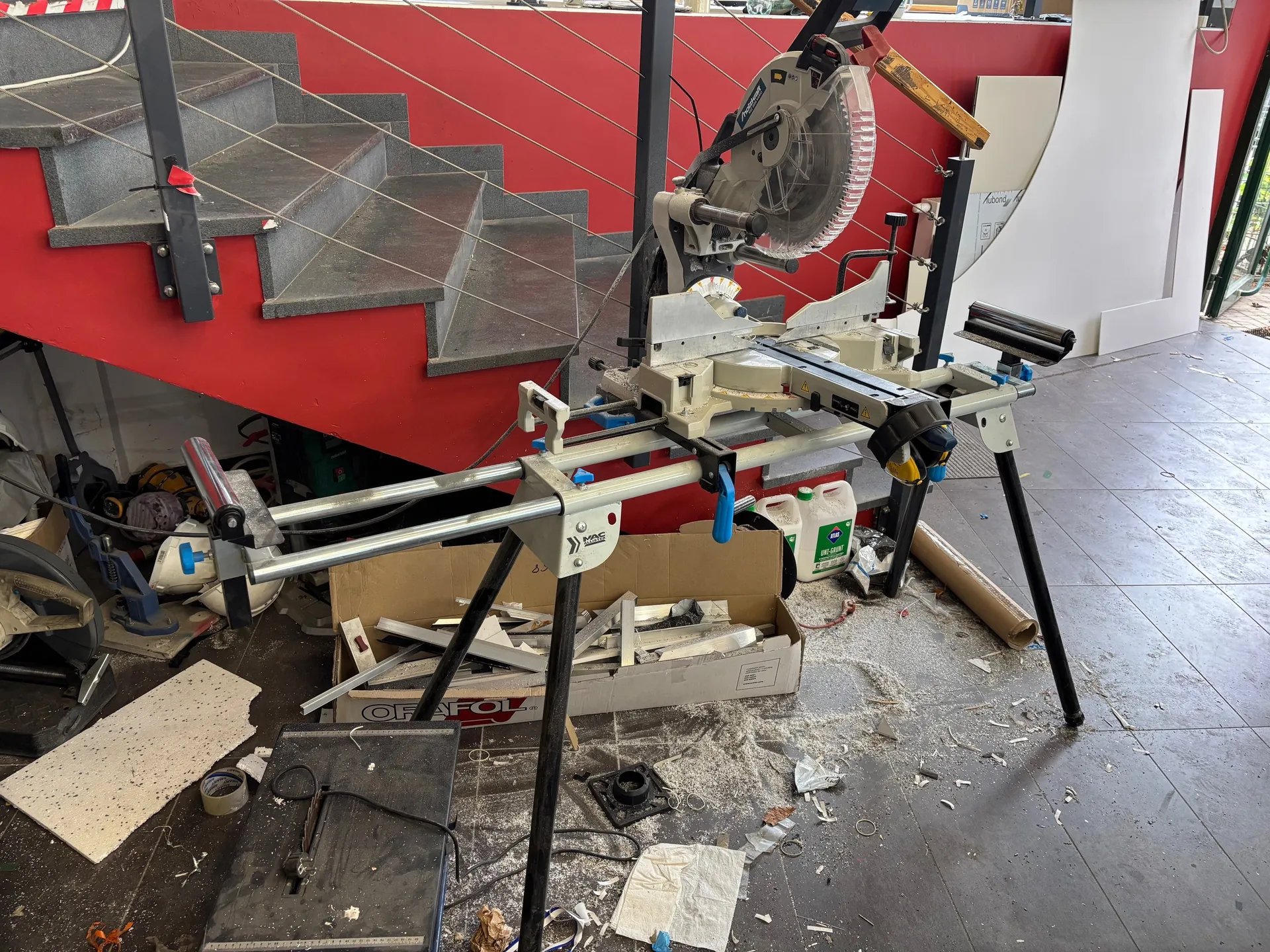 Pracownia z maszyną do cięcia pod kątem (miter saw) na statywie z czarnymi nogami. W tle czerwona klatka schodowa z szarymi stopniami i metalowymi poręczami z linami. Na podłodze rozrzucone narzędzia, pudełko z napisem ORAFOL wypełnione metalowymi elementami, białe pojemniki z zielonymi etykietami, rola brązowego materiału oraz śmieci i wióry. Czerwona ściana i biała konstrukcja po prawej stronie.