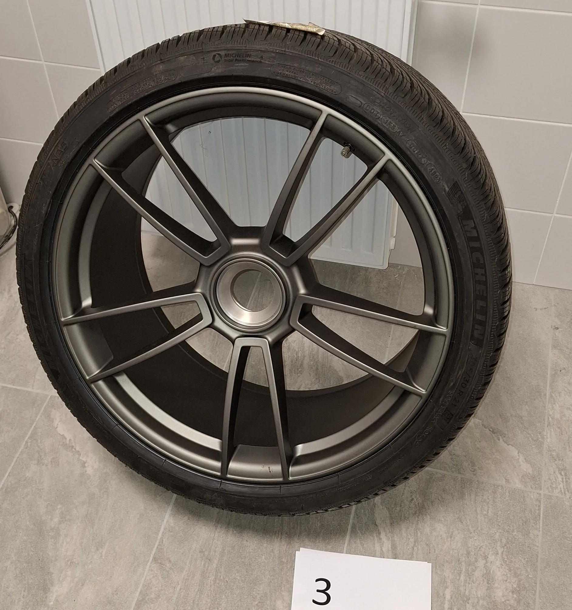 Zdjęcie koła samochodowego z oponą marki Michelin i ciemnoszarą, wieloramienną felgą aluminiową. Koło opiera się o jasną, kafelkową ścianę i podłogę. Na dole obrazu widoczna jest biała kartka z czarną cyfrą 3.