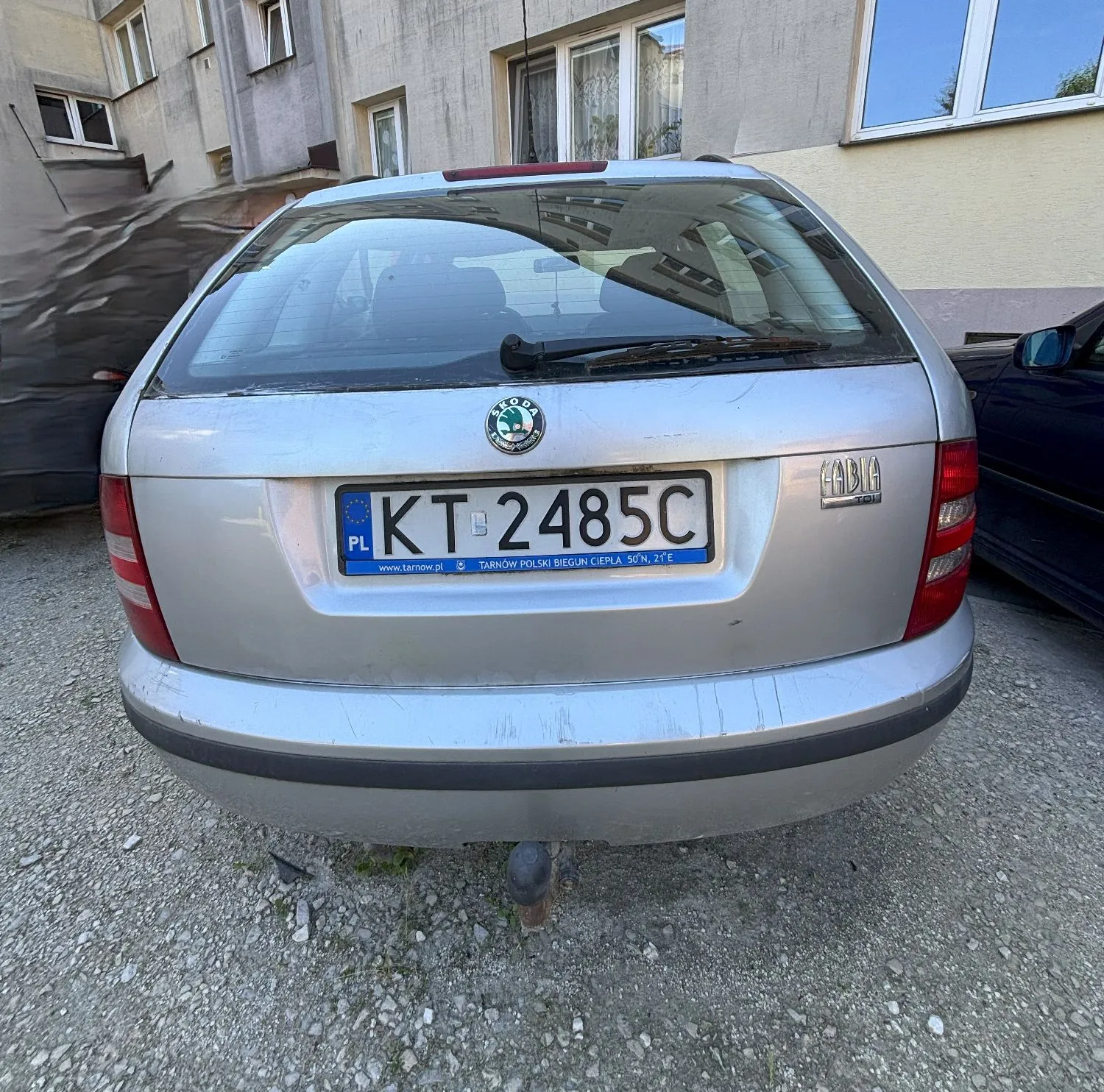 Tylny widok srebrnego samochodu marki Skoda Fabia Combi. Pojazd posiada polską tablicę rejestracyjną o numerze KT 2485C z napisem 'TARNÓW POLSKI BIEGUN CIEPŁA 50°N, 21°E' na ramce. Samochód jest wyposażony w hak holowniczy i zaparkowany na żwirowym podłożu przed budynkiem mieszkalnym.