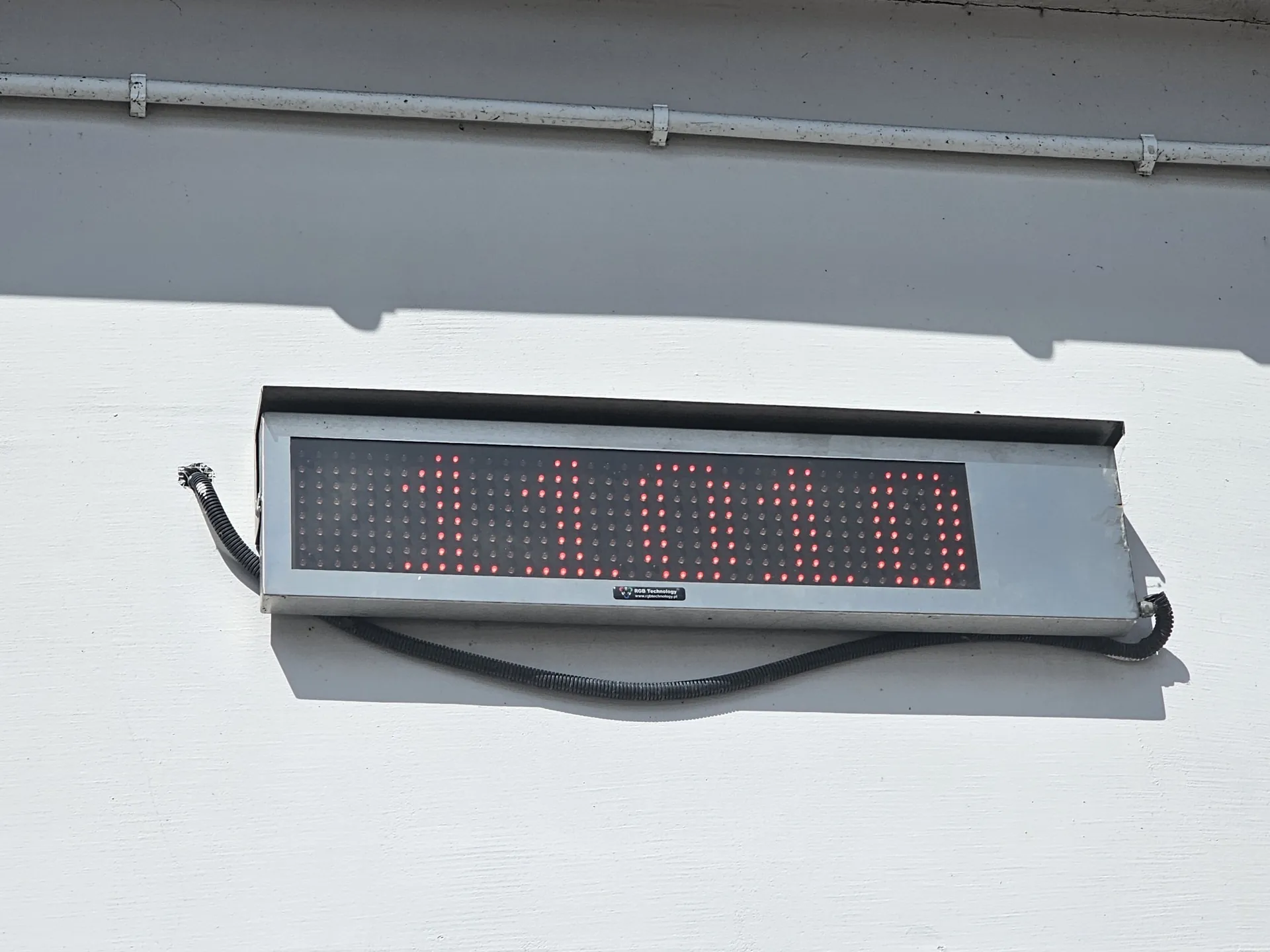 Elektroniczny wyświetlacz LED zamontowany na białej ścianie. Wyświetlacz ma srebrną obudowę i pokazuje czerwone cyfry, które są częściowo nieczytelne z powodu rozmycia ruchu, przypominając liczbę 1010. Od spodu urządzenia odchodzi czarny, karbowany przewód elektryczny. Nad wyświetlaczem przebiega pozioma, biała rura zamocowana do ściany obejmami.