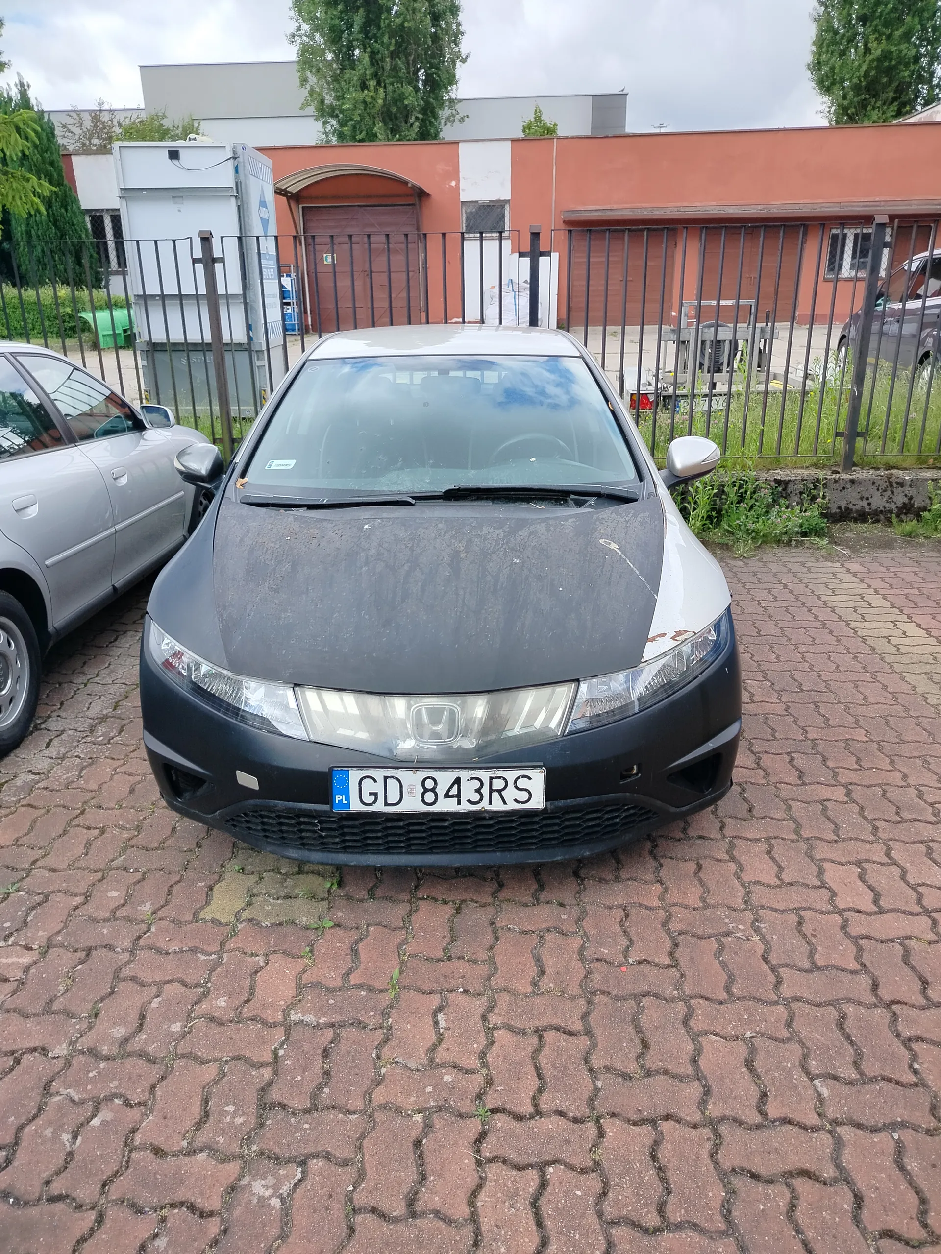 Czarny samochód marki Honda z tablicą rejestracyjną GD 843RS (PL) zaparkowany na ceglanym placu. Po lewej stronie częściowo widoczny srebrny samochód. W tle metalowe ogrodzenie, pomarańczowo-białe budynki, brama garażowa oraz drzewa. Niebo jest zachmurzone.