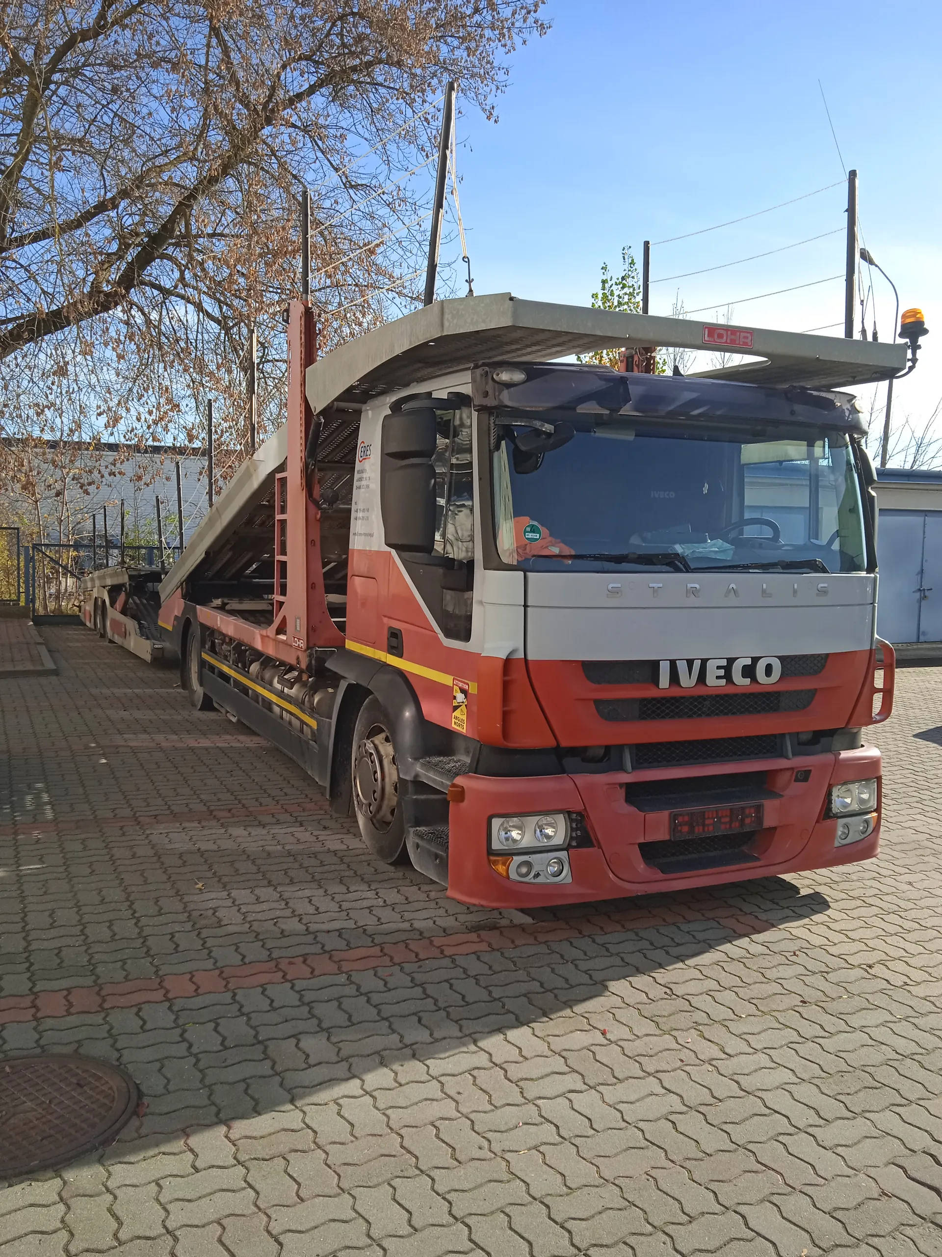 Czerwono-biały ciężarówka Iveco Stralis z przyczepą do przewozu samochodów marki LOHR, zaparkowana na brukowanej powierzchni. Na przodzie widoczne napisy 'IVECO' i 'STRALIS', a na przyczepie czerwony napis 'LOHR'. W tle drzewa, budynki i jasne niebo. Tekst na obiekcie jest czytelny i ustawiony pionowo.