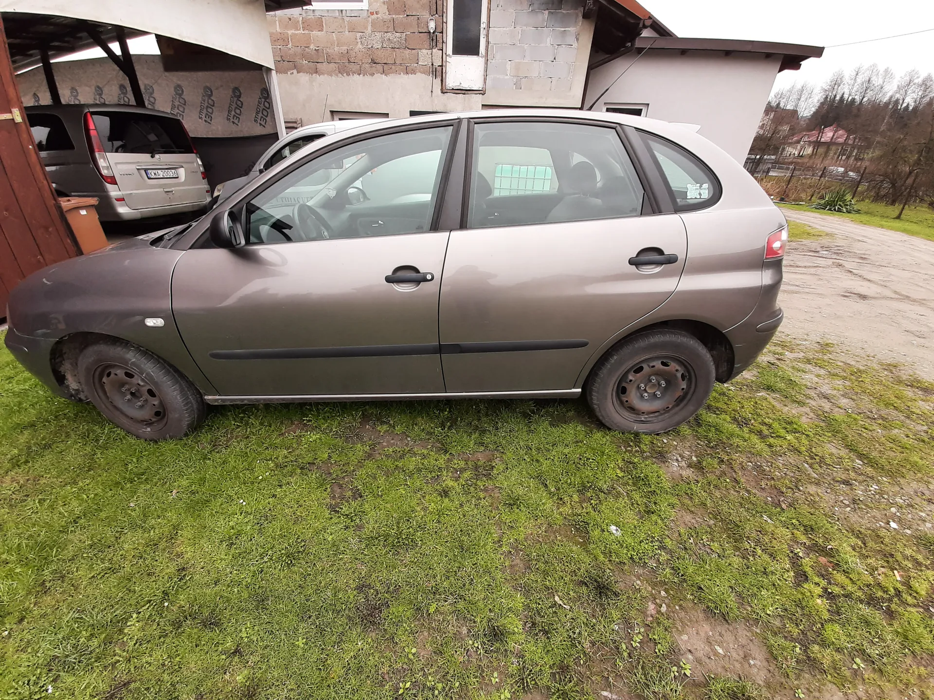 Szary samochód osobowy typu hatchback zaparkowany na trawie, widok z boku. W tle garaż z częściowo widocznym srebrnym samochodem o tablicy rejestracyjnej KMA 20034. Budynki z cegły i betonu, zielona trawa na pierwszym planie, droga ziemna po prawej stronie. W oddali drzewa i domy.