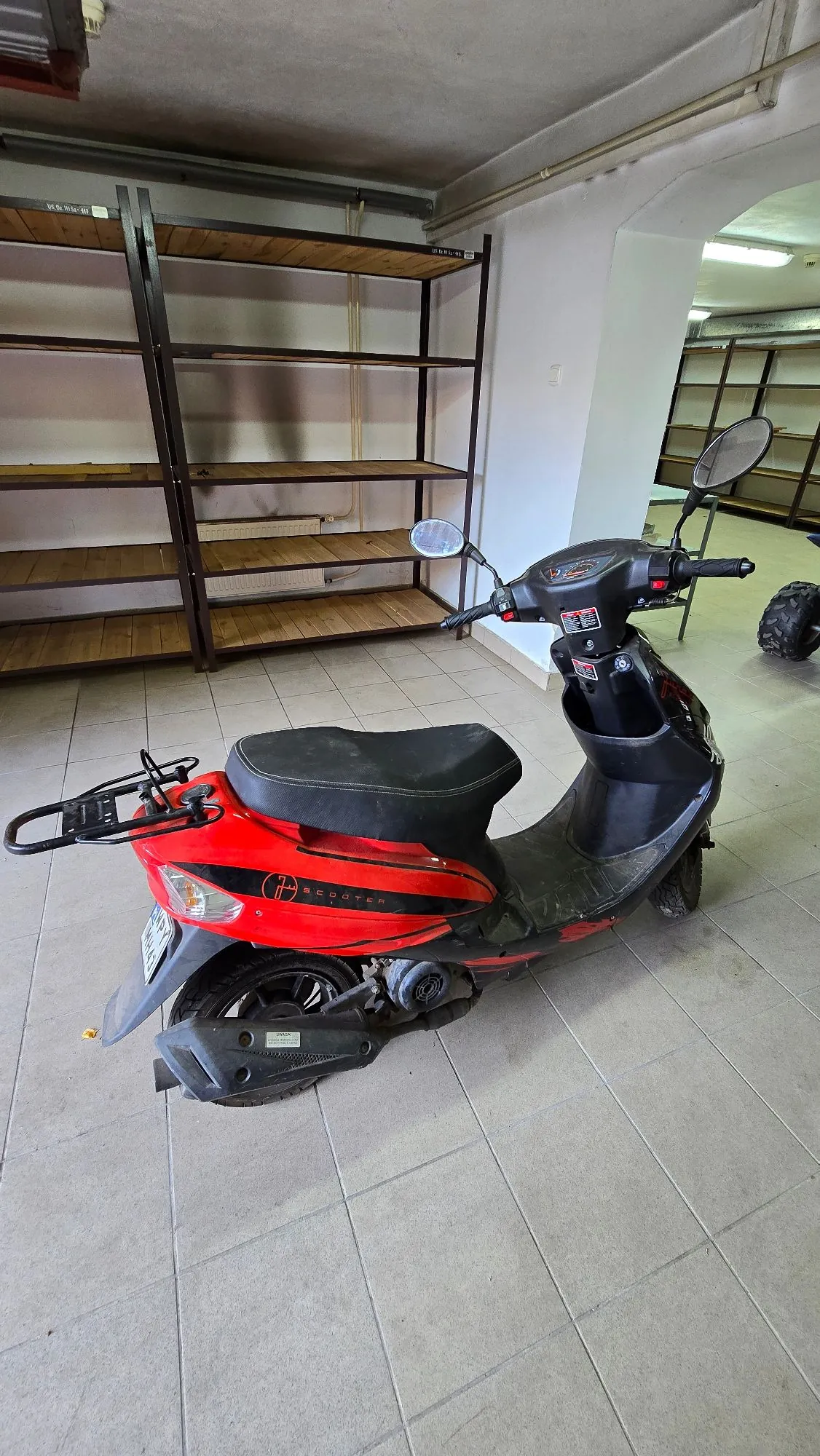 Czerwono-czarny skuter zaparkowany w pomieszczeniu z jasną kafelkową podłogą. W tle widoczny jest duży, drewniany regał z metalową konstrukcją oraz biała ściana z łukowatym przejściem do innego pomieszczenia.