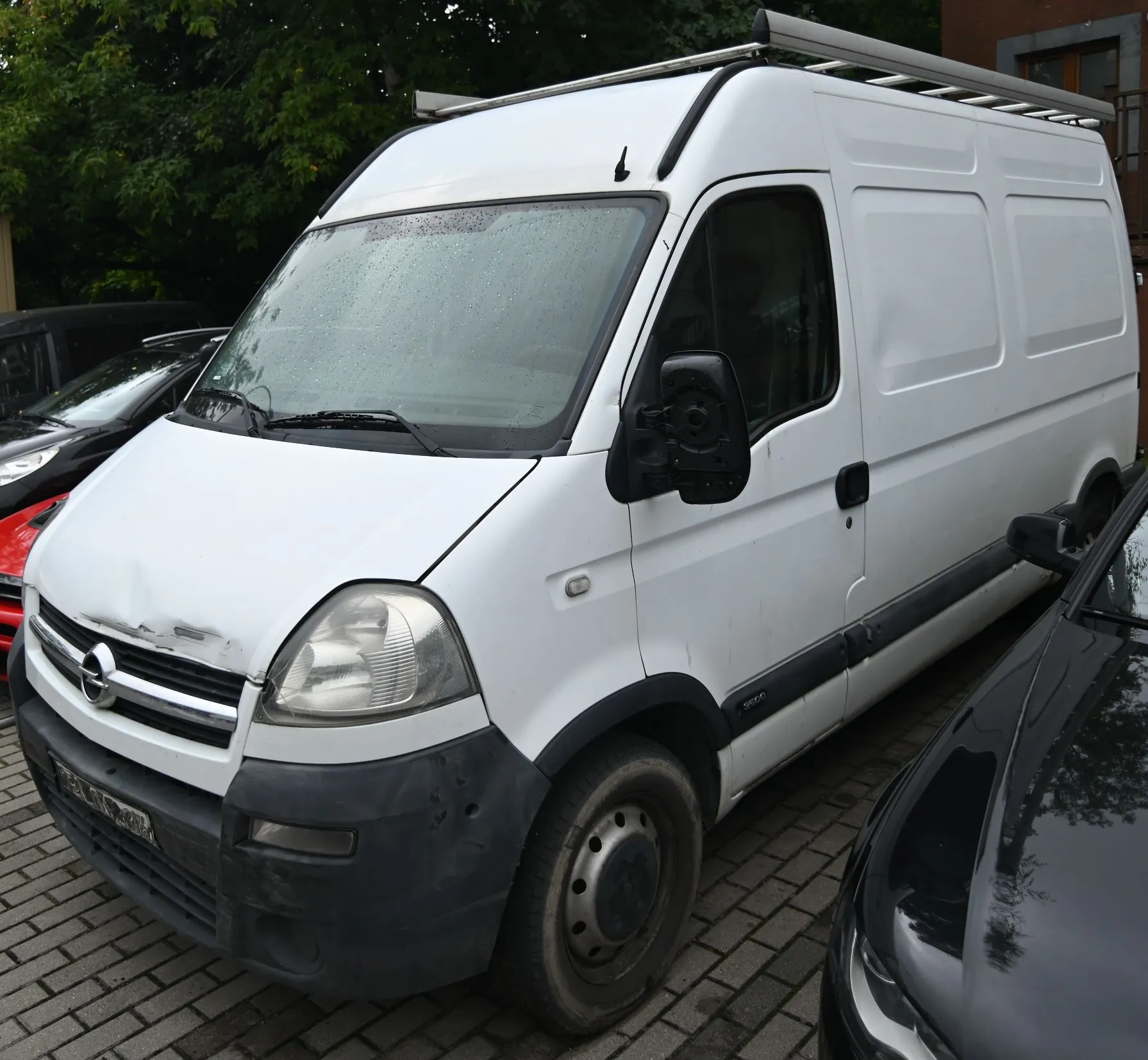 Biały furgon marki Opel Vivaro zaparkowany na kostce brukowej między dwoma czarnymi samochodami. Pojazd posiada bagażnik dachowy, a na masce widoczne jest niewielkie wgniecenie nad grillem.